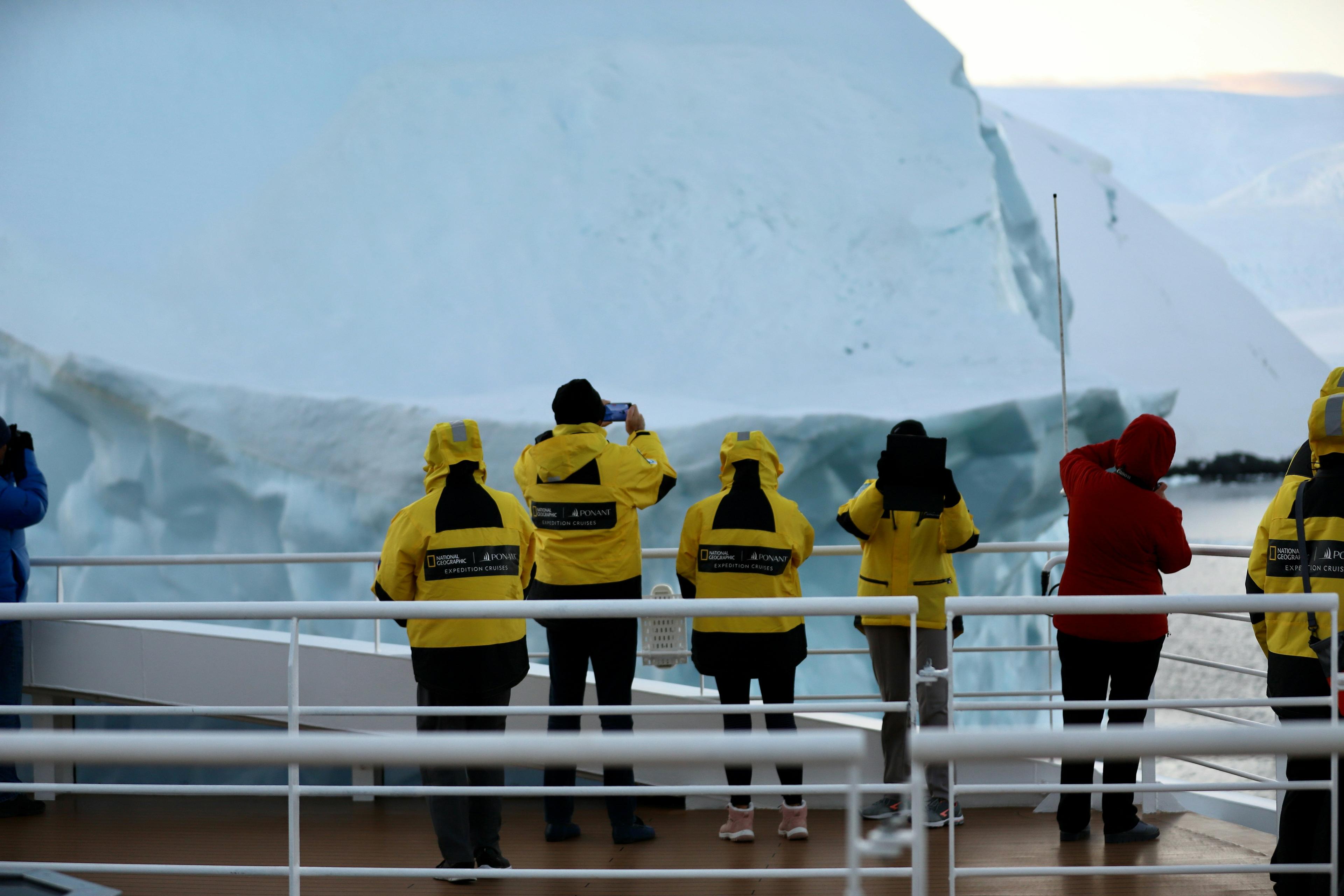 Passengers wearing yellow jackets capture photographs of a massive iceberg from the deck of a National Geographic expedition cruise ship, likely in Antarctic waters.