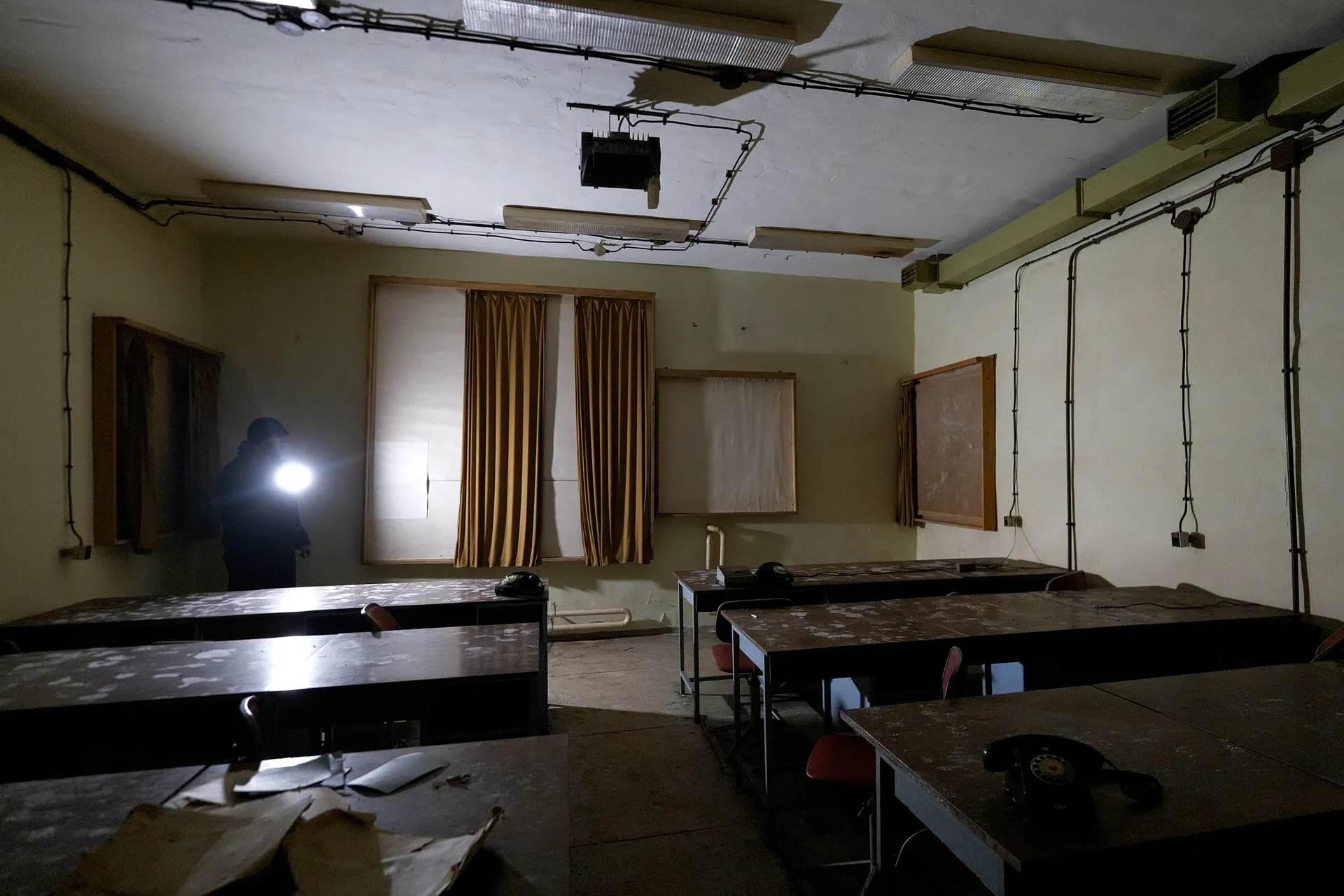 A dimly lit abandoned bunker room, where a faint light breaks through the heavy darkness. Simon stands setting up camera gear amidst rows of decaying desks covered in white mold