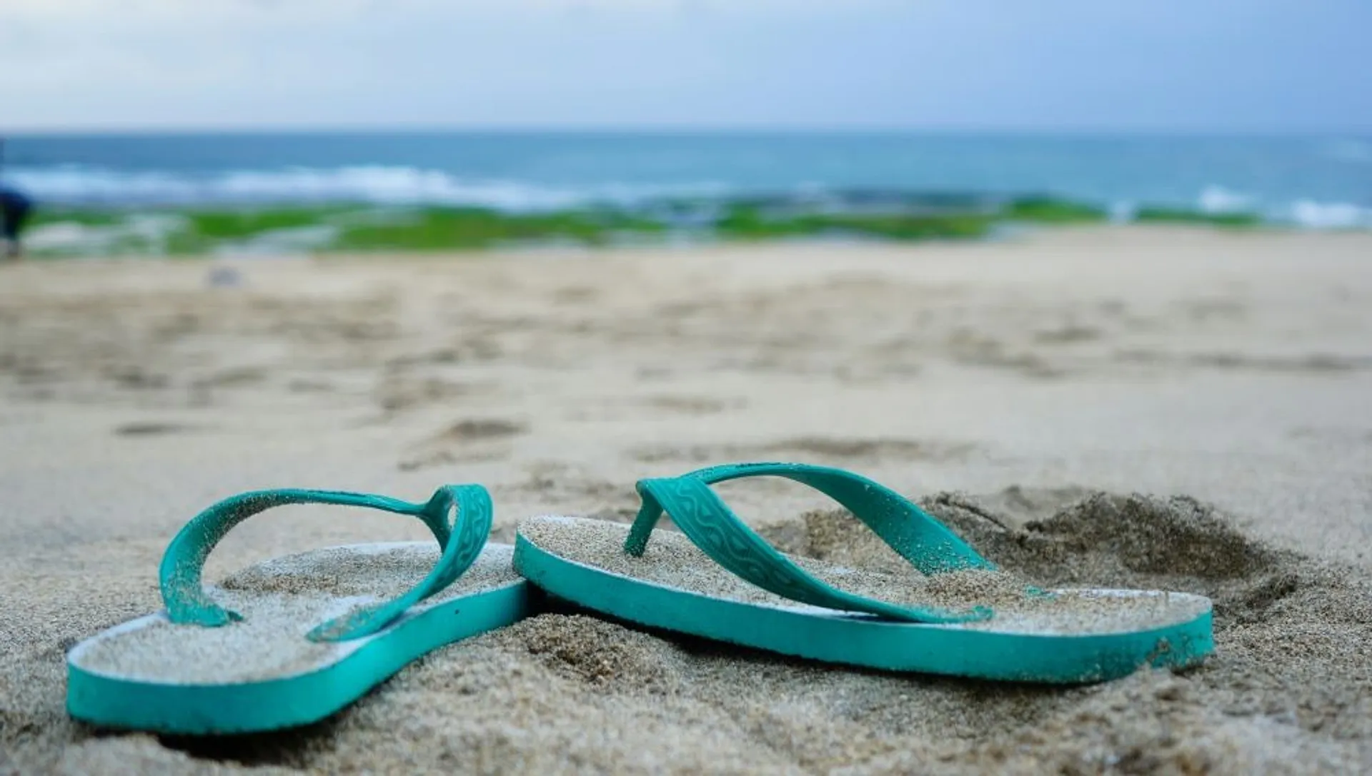 Turquoise flip-flops rest in the sand on a tranquil beach with gentle waves in the background, possibly reminiscent of a tropical getaway like Bali.