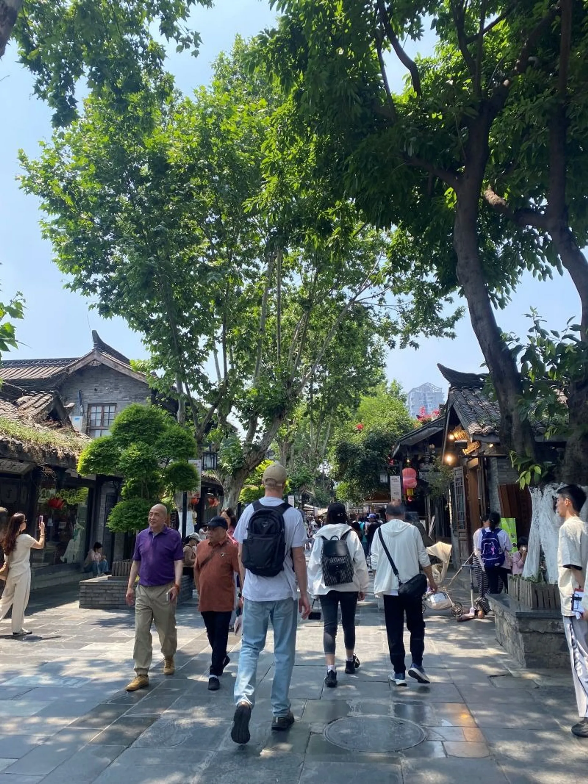 Pedestrians stroll through a lively, tree-lined street in Chengdu's Jinli Ancient Street, surrounded by historic-style buildings.
