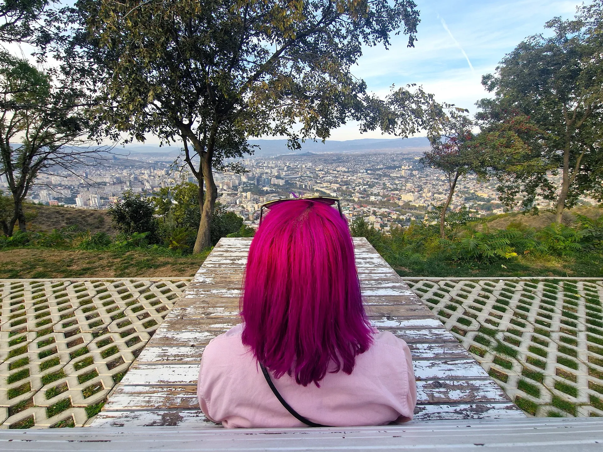 A person with vibrant pink hair sits on a bench overlooking the sprawling cityscape of Tbilisi, Georgia from a scenic vantage point.