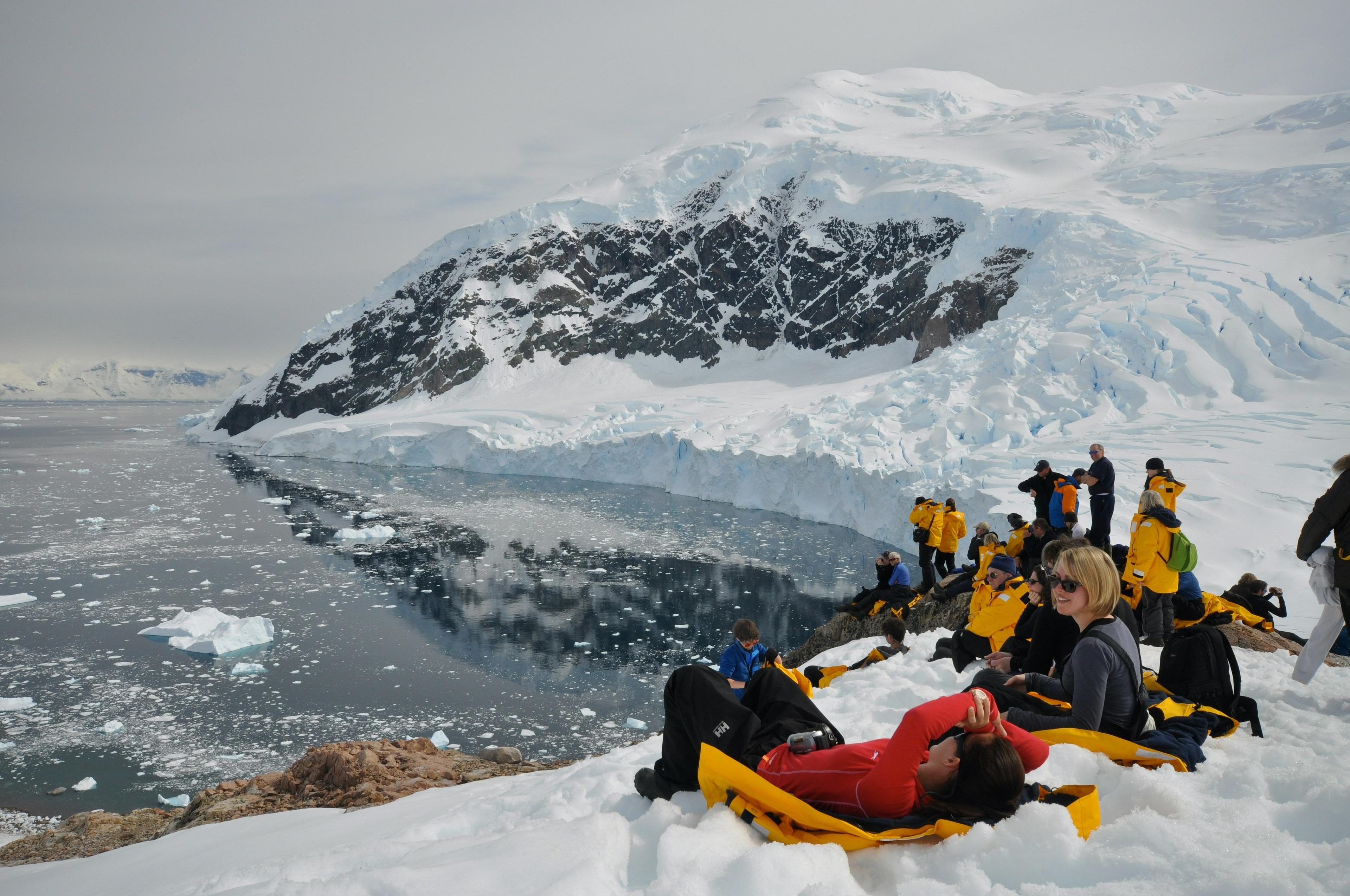 A group of people dressed warmly in vibrant clothing relax and explore the icy landscape near Neko Harbour, Antarctica, with snow-covered mountains and ice-filled waters stretching into the distance.