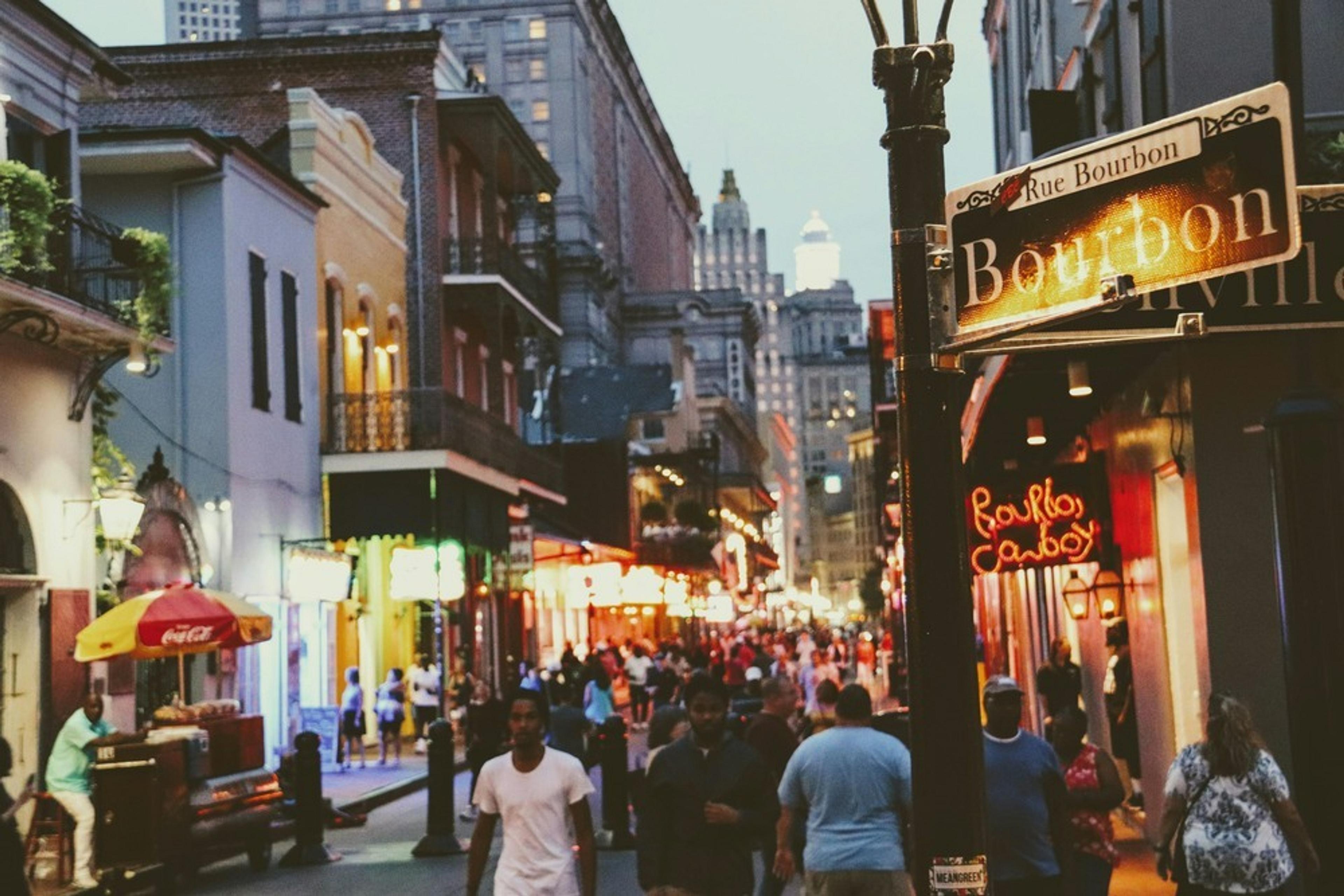 A bustling evening scene unfolds on Bourbon Street in New Orleans, with neon lights illuminating the vibrant crowds.