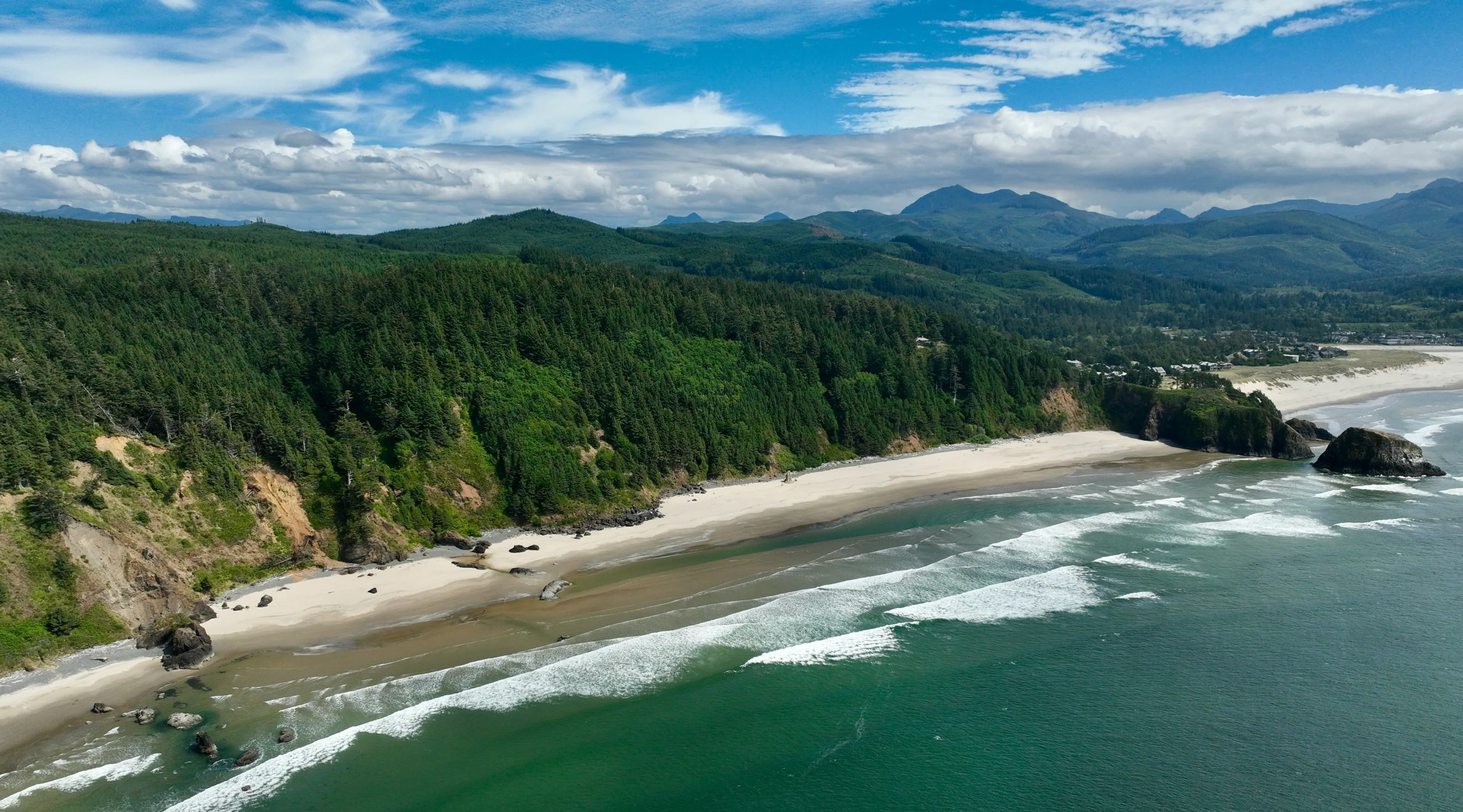 Waves gently crash along the sandy shores of Cannon Beach with lush forests and distant mountains as a backdrop.