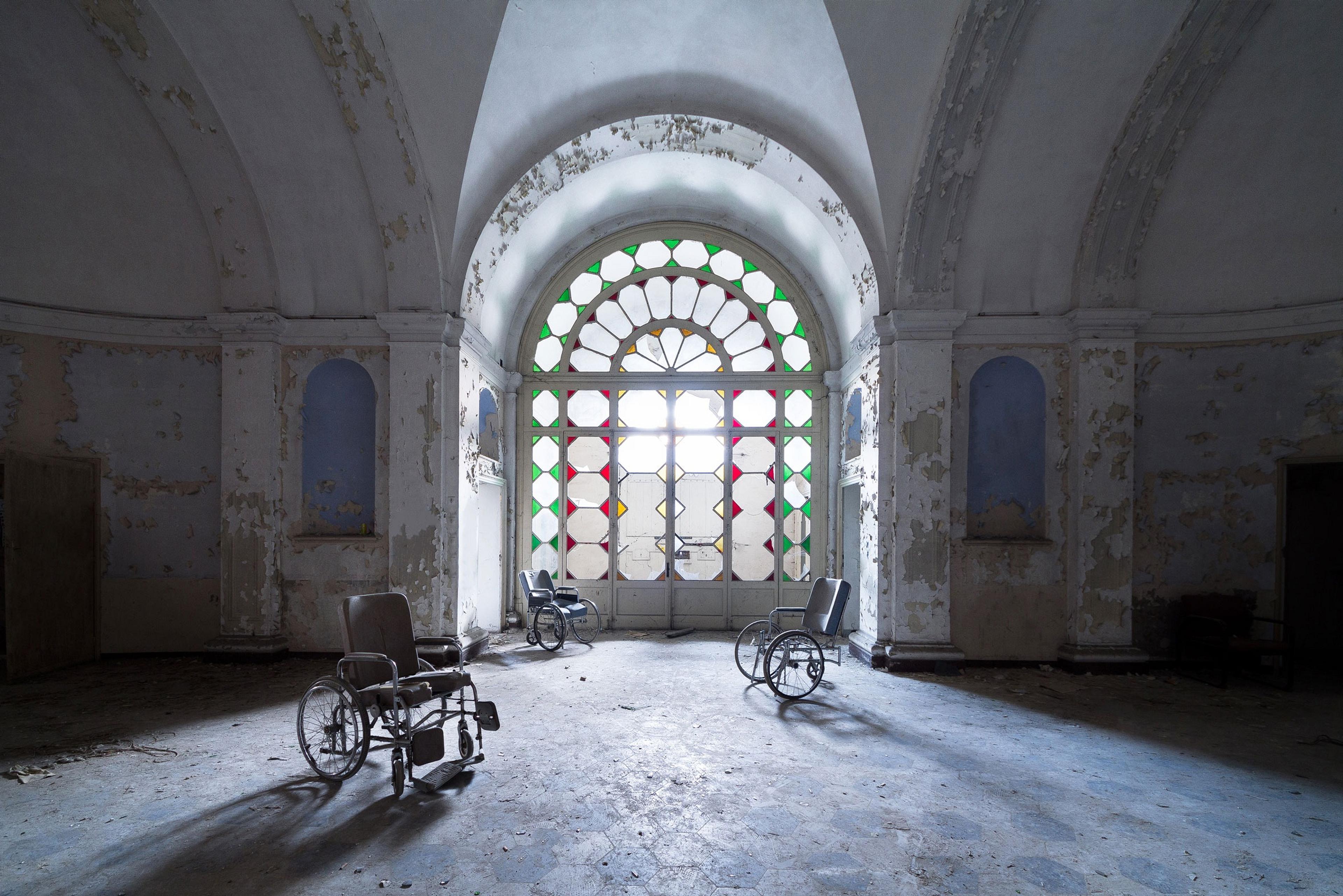 An abandoned Italian psychiatric asylum's grand hall, where sunlight filters through a stained glass window, casting light across a lone wheelchair. 