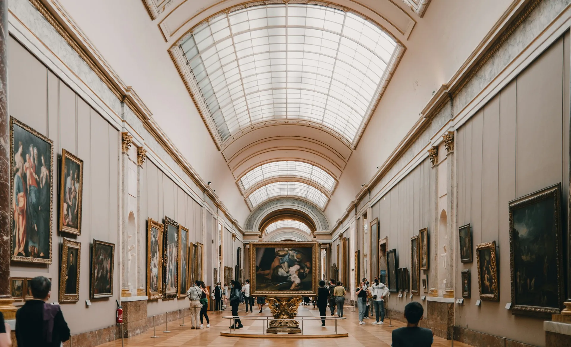 Visitors admire artwork in the gallery of the Louvre Museum in Paris, under a grand arched ceiling.