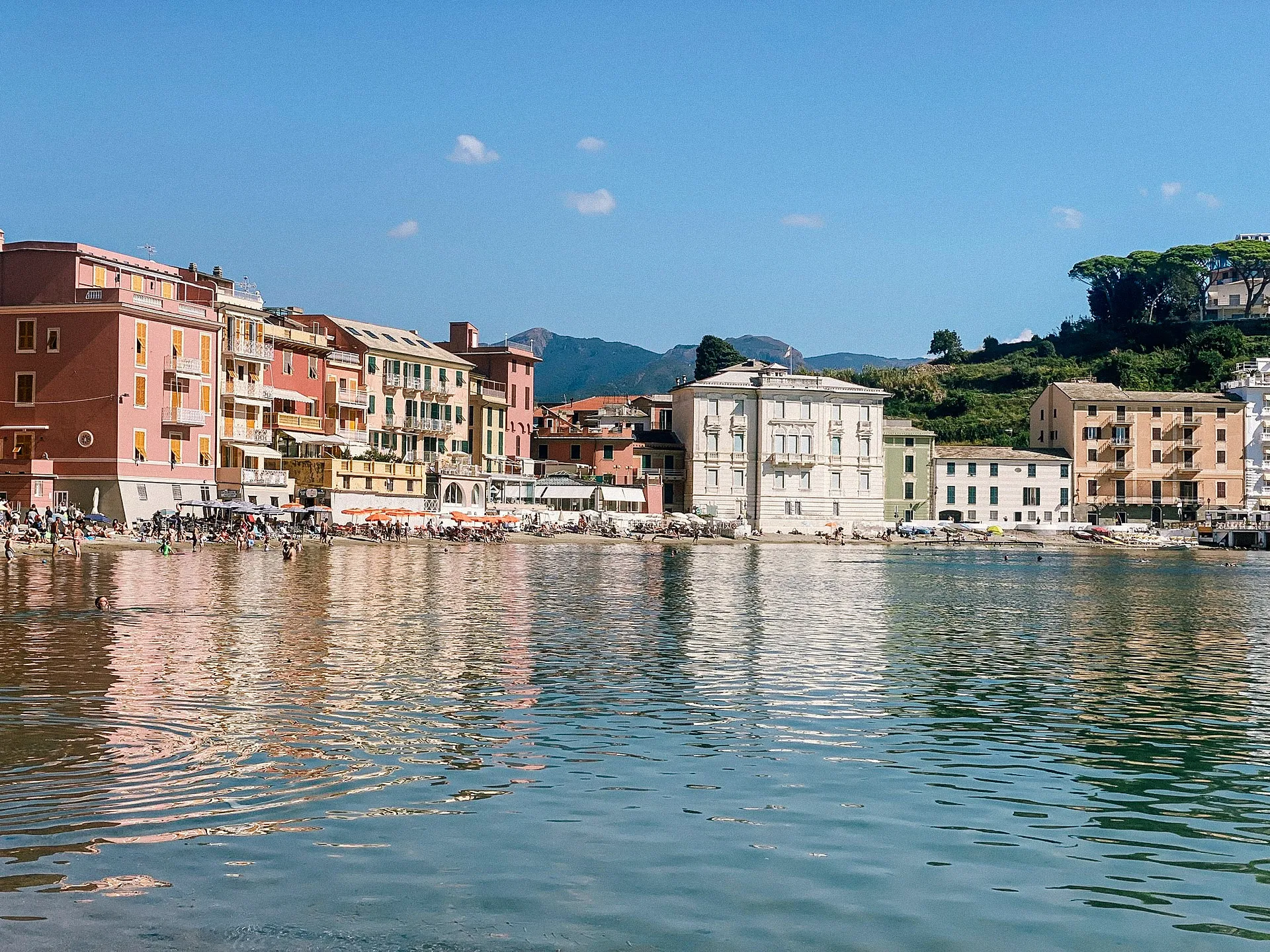 A vibrant seaside scene at Bay of Silence in Sestri Levante, Italy, features colorful buildings and sunbathers along the shoreline.