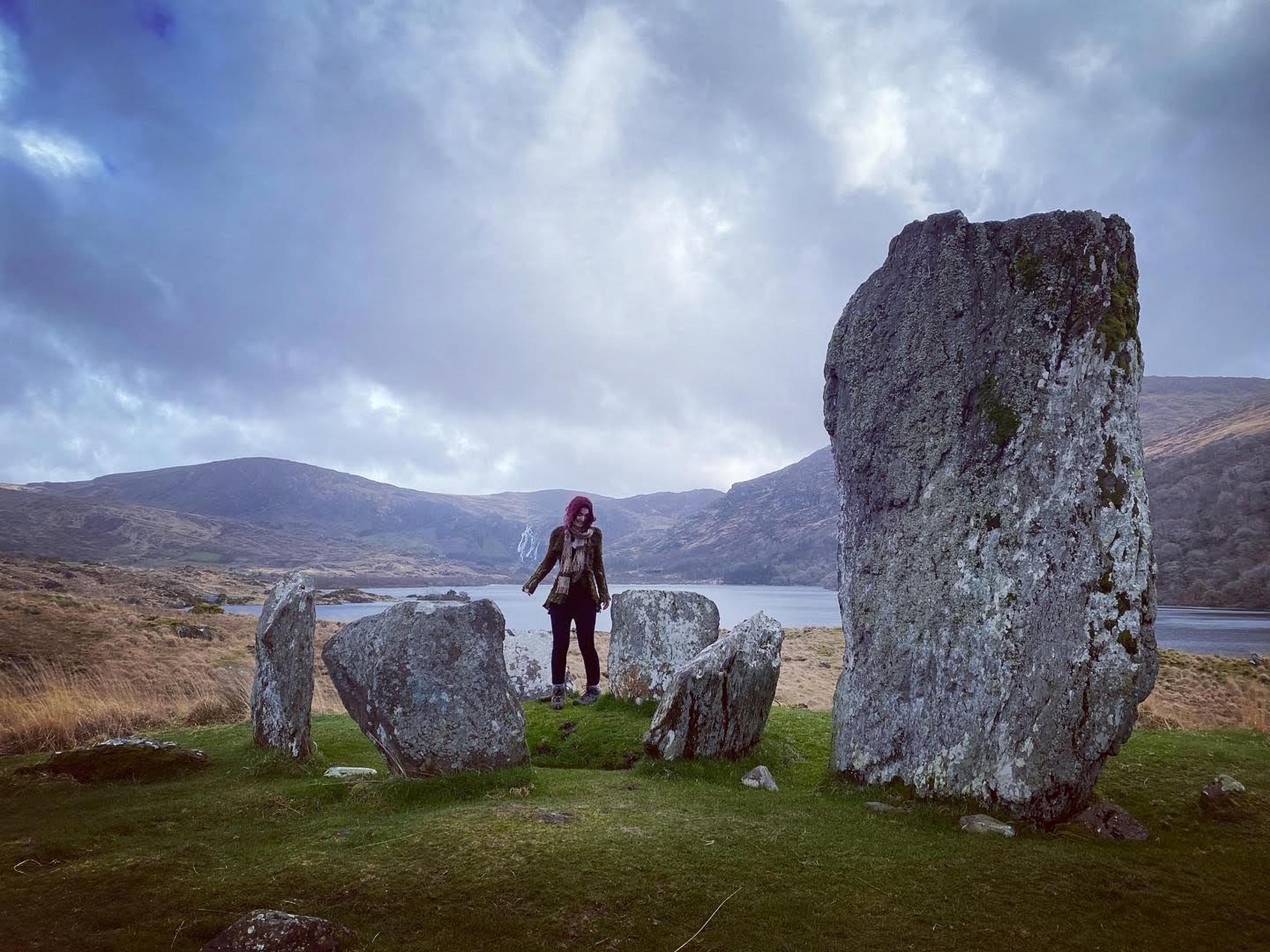 Akasha standing in a circle of stones, mountains and water in the distance