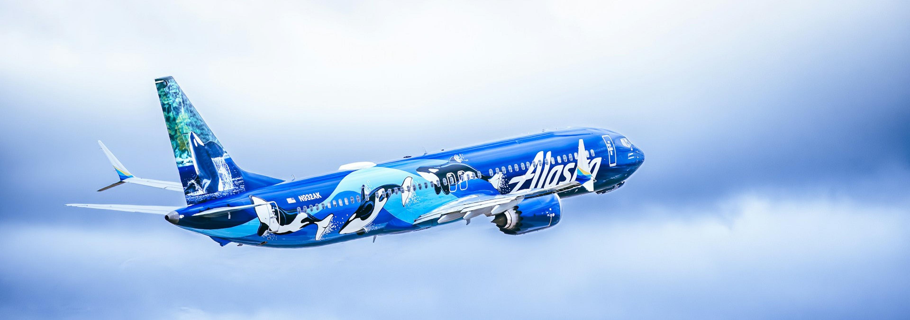 An Alaska Airlines plane featuring orca designs soars through a cloudy sky.
