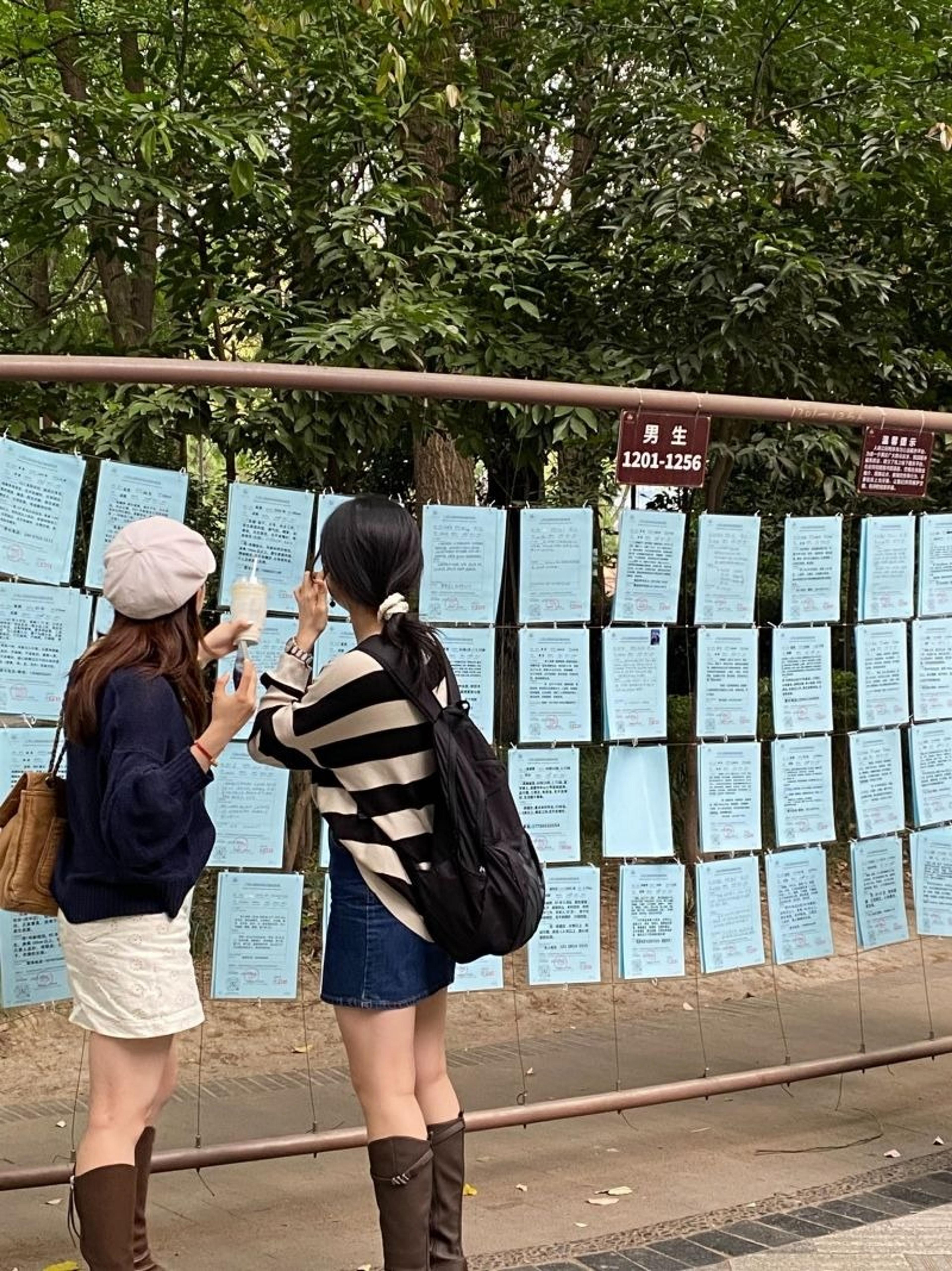 Two people examine handwritten notes on a bulletin board in Chengdu's People's Park