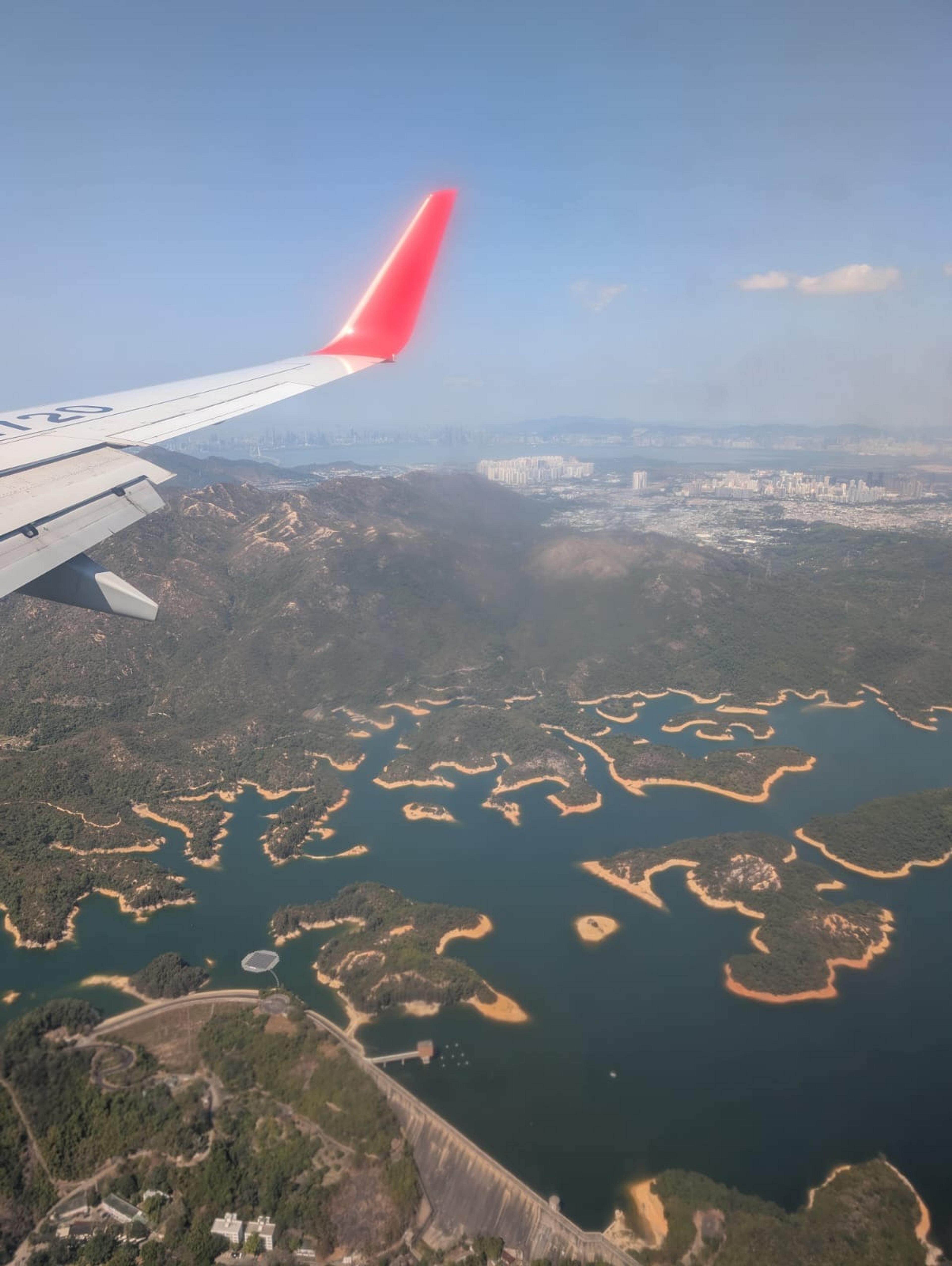 An aerial view of the Plover Cove Reservoir surrounded by lush hills and the cityscape of Hong Kong in the distance.