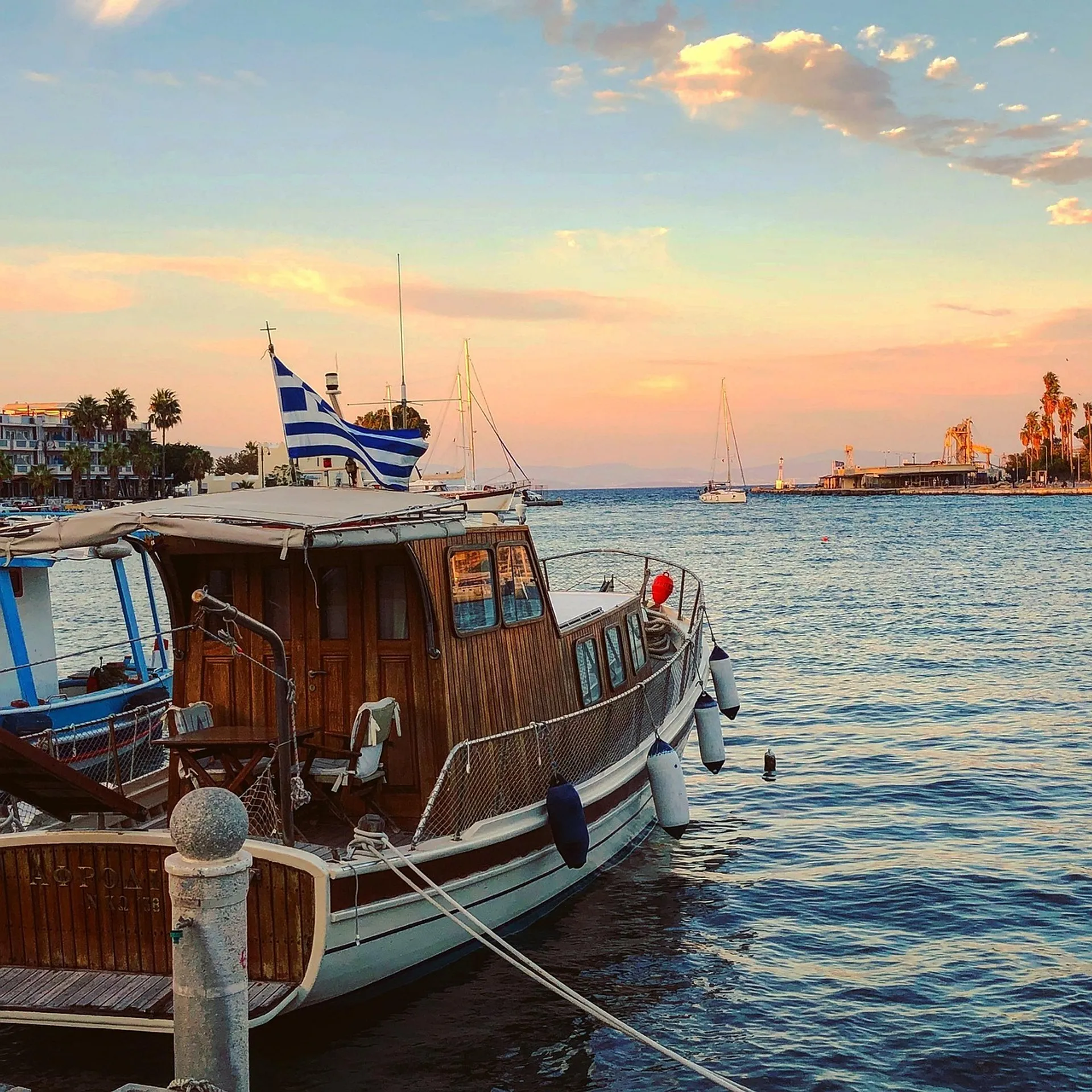 A wooden boat with a Greek flag is docked at the harbor in Kos Town, Greece, during a colorful sunset.