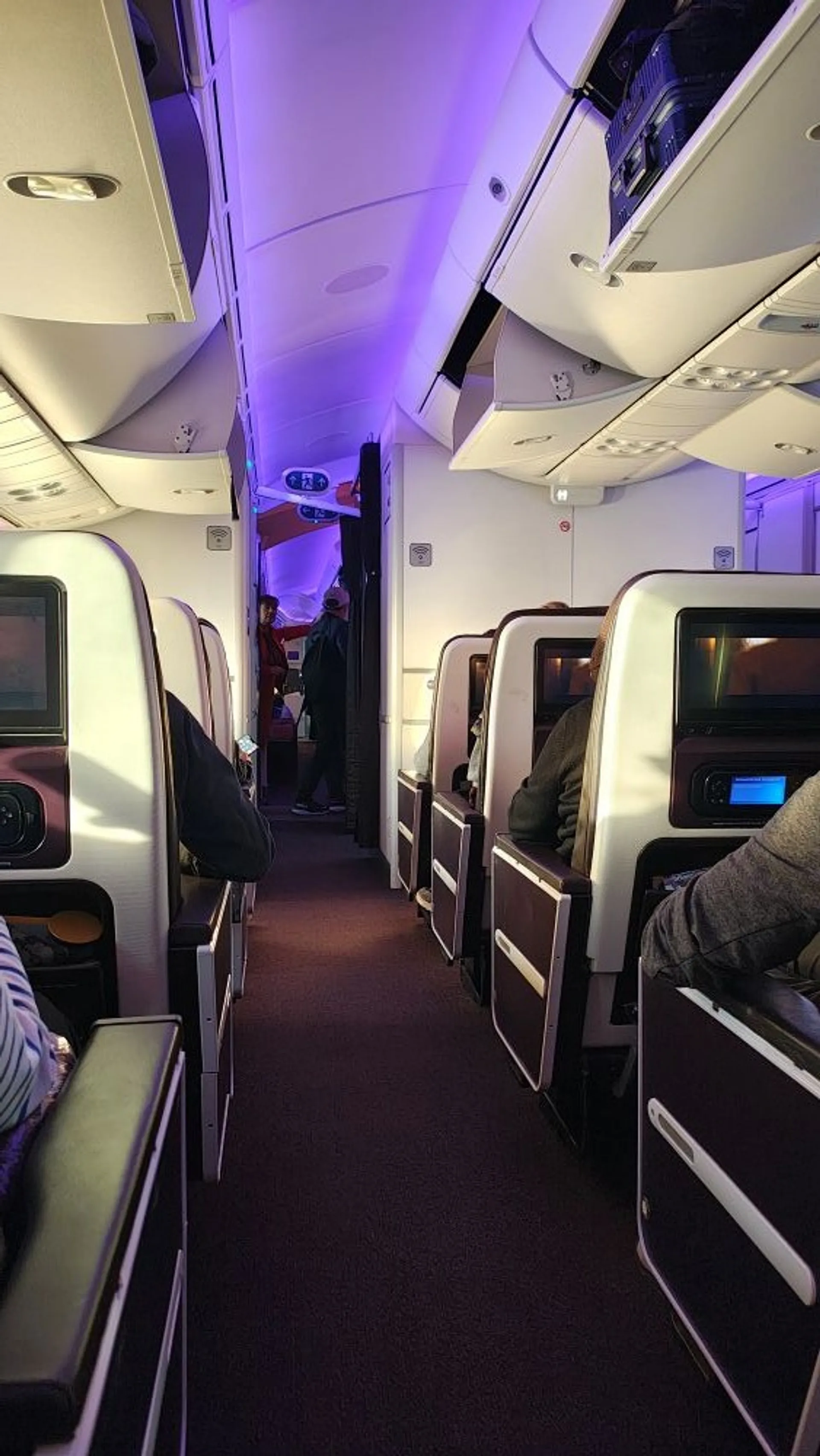 A view down the aisle of a Virgin Atlantic aircraft cabin with blue and purple lighting and passengers seated.