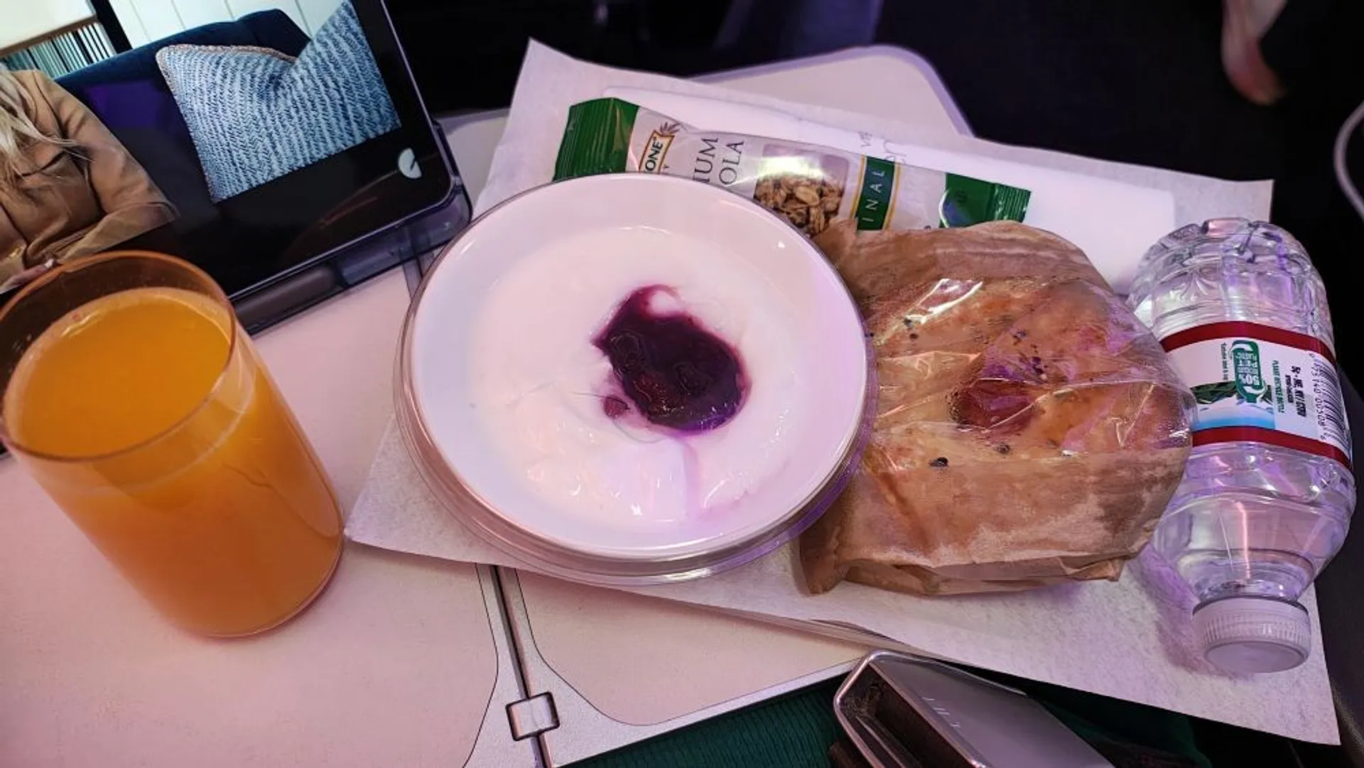 A breakfast consisting of yogurt, an orange drink, packaged pastry, and bottled water is arranged on an airplane tray table.