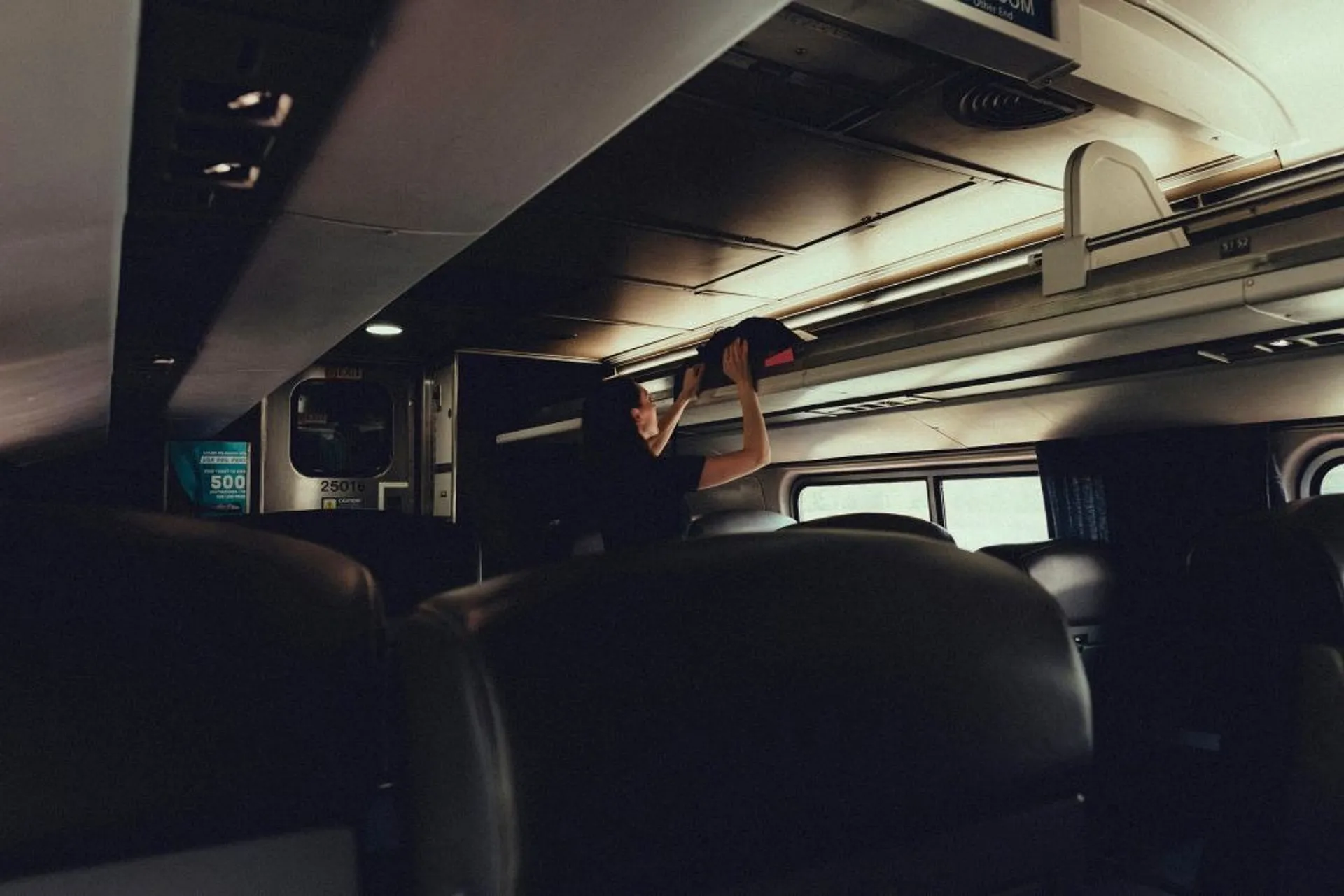 A person places luggage in the overhead compartment inside an Amtrak train, with car number 25018 visible.