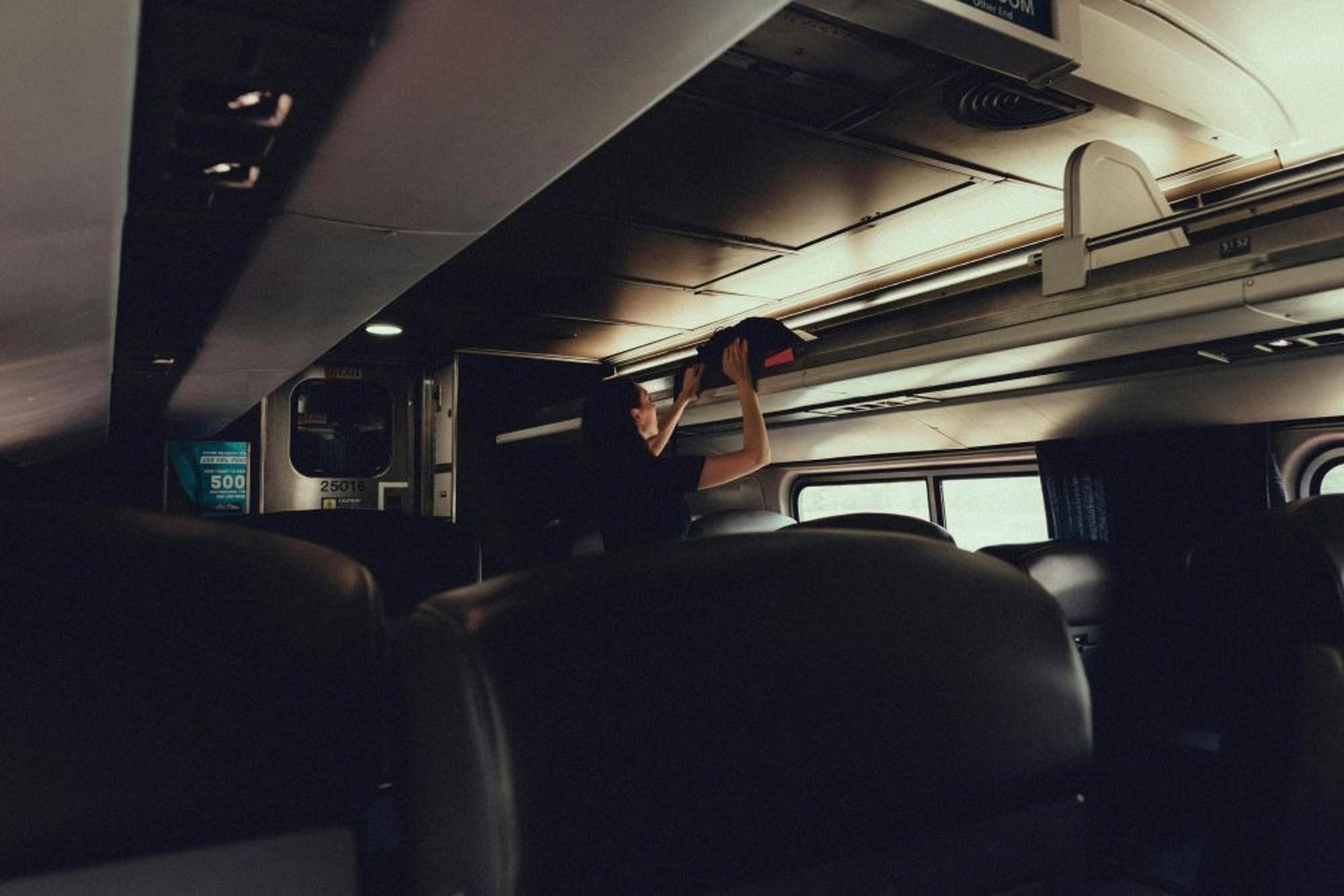 A person places luggage in the overhead compartment inside an Amtrak train, with car number 25018 visible.