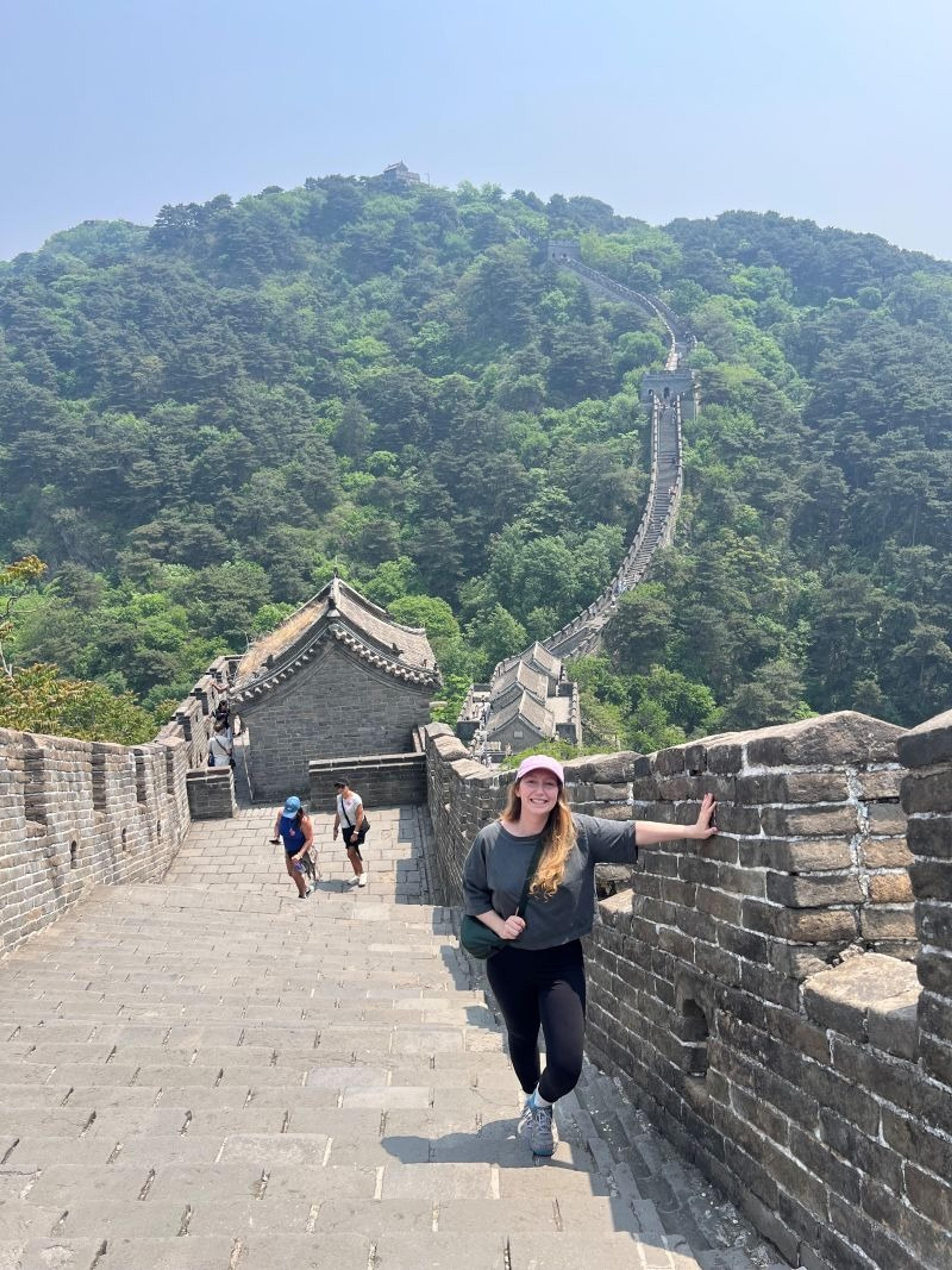 A woman smiles while walking along the Great Wall of China amidst lush, green hills.