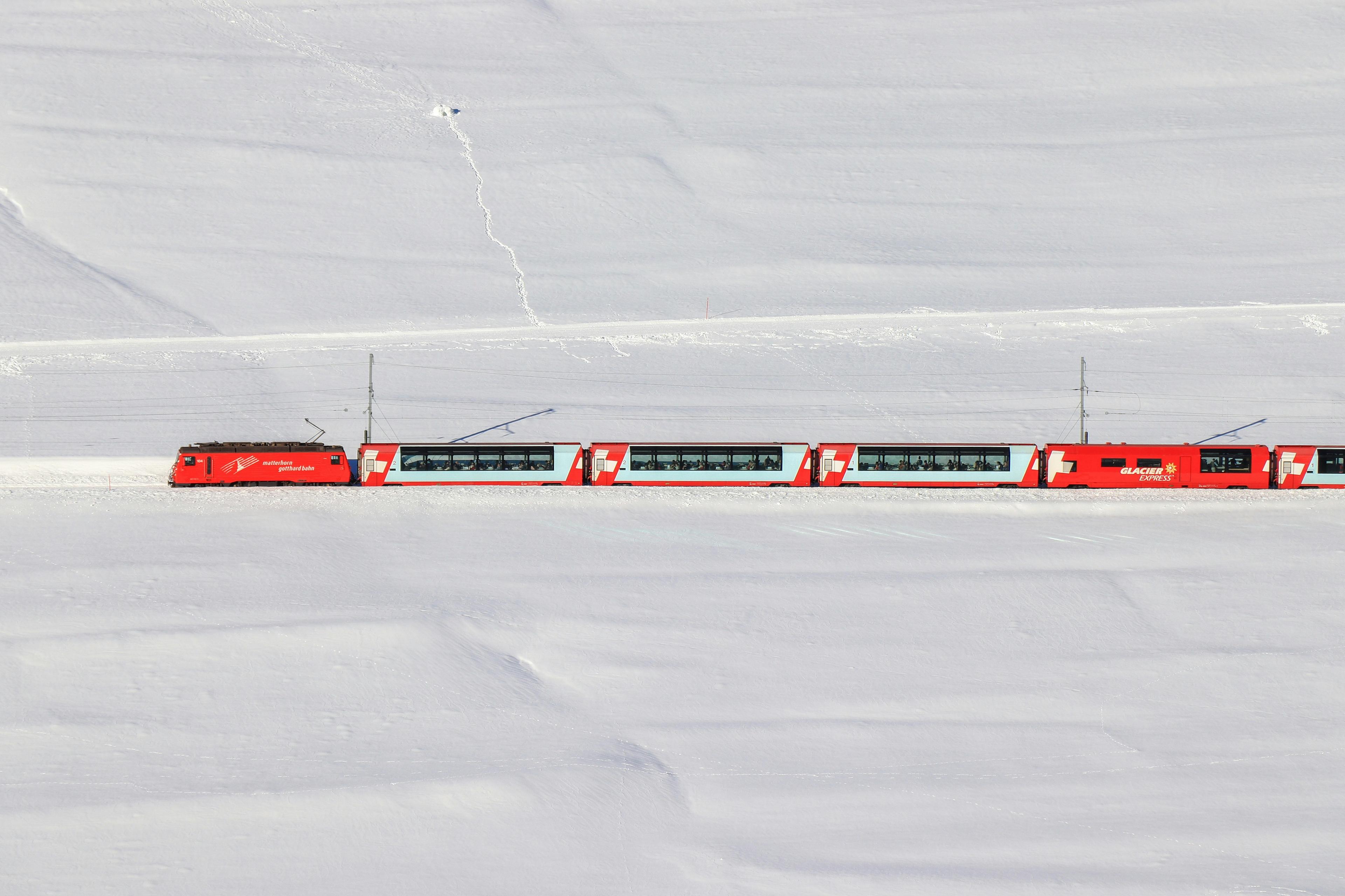 A bright red Glacier Express train travels across the snowy landscape of the Swiss Alps.