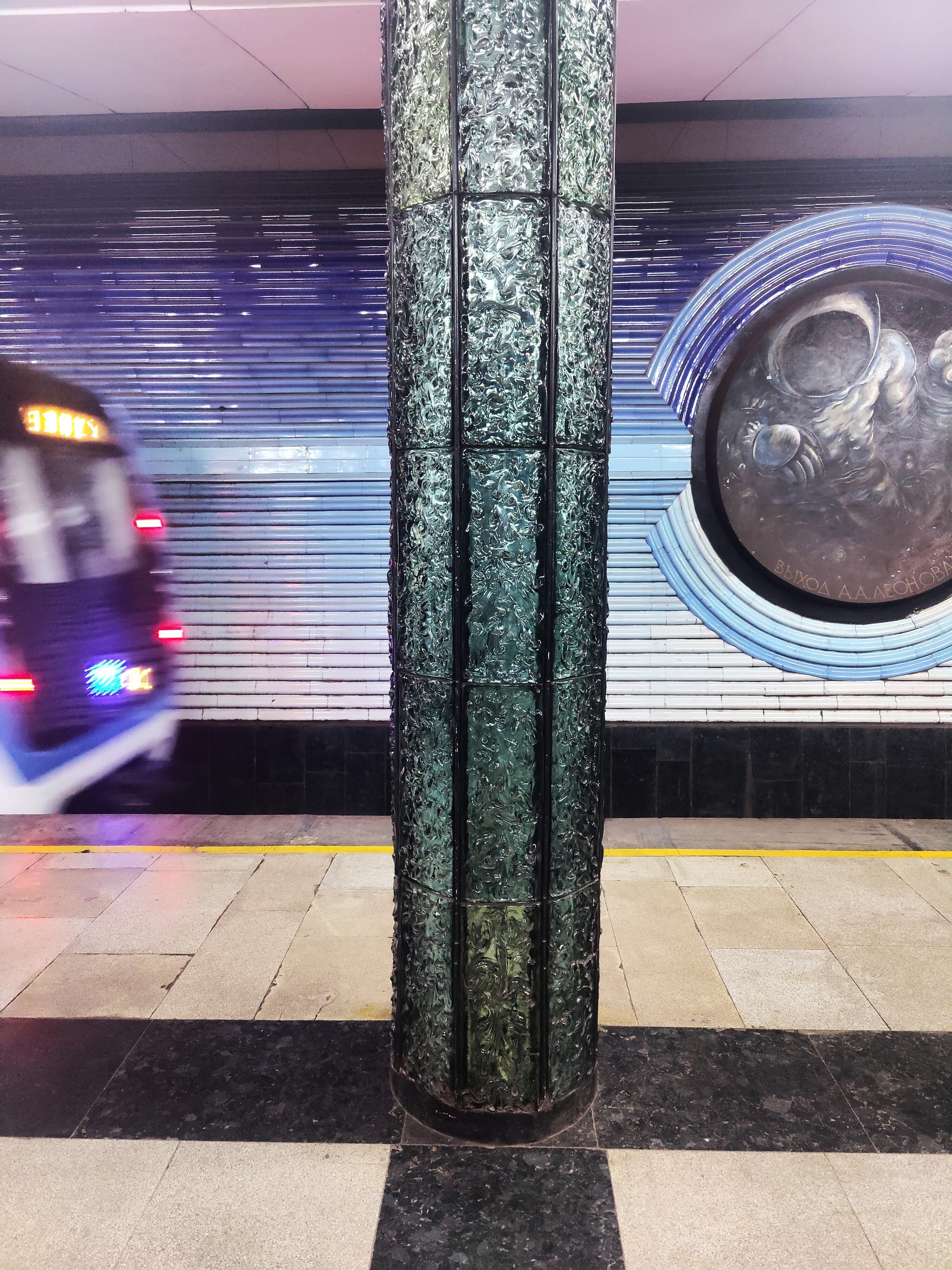 A train speeds past a uniquely designed column at Elektrozavodskaya metro station in Moscow, featuring blue and white tiled walls and a circular relief.