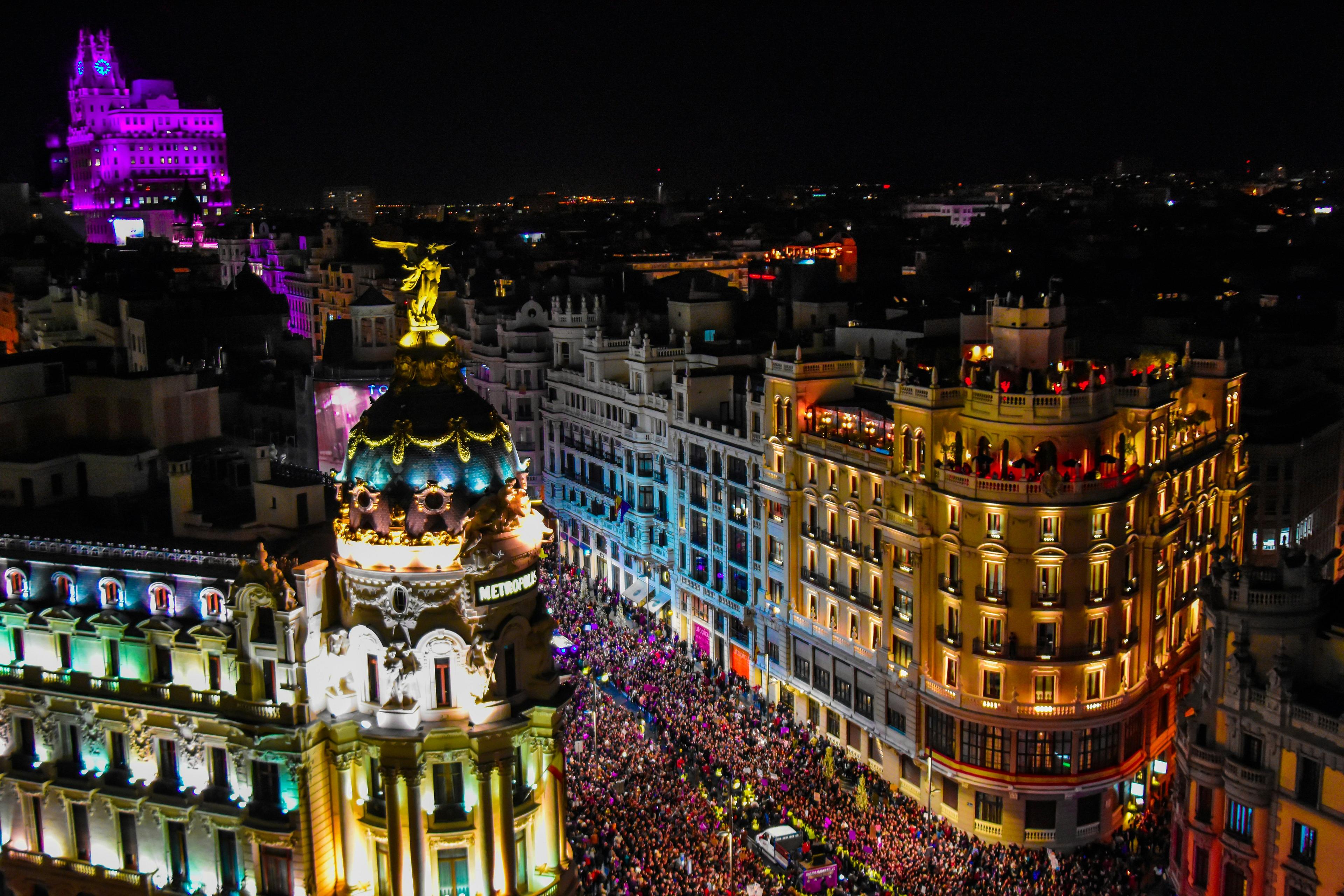A vibrant nighttime scene captures the bustling Gran Vía in Madrid, with the iconic Metropolis Building illuminated and crowds filling the street.