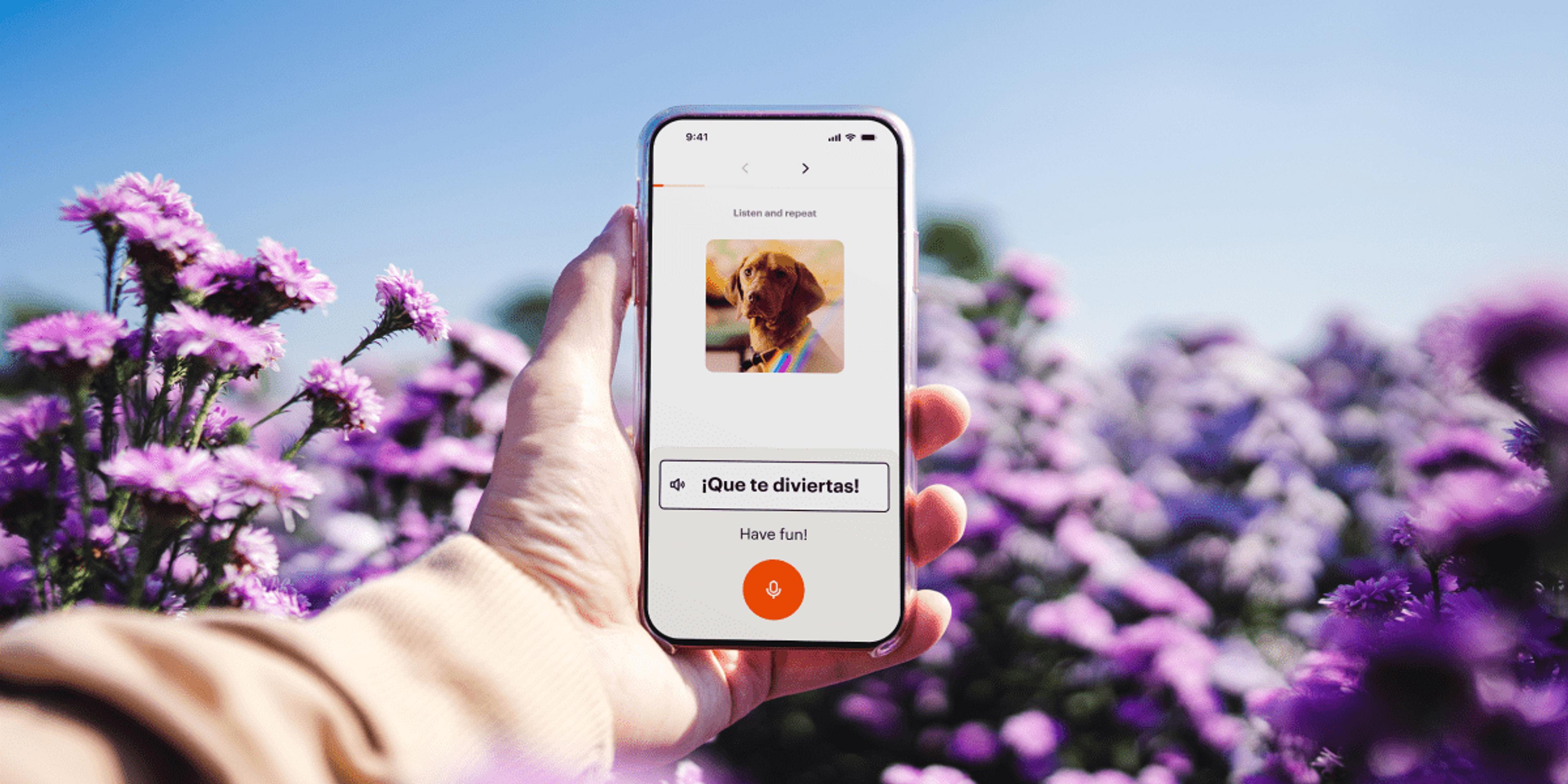 A hand holds a smartphone displaying a language learning app in a field of vibrant purple flowers.