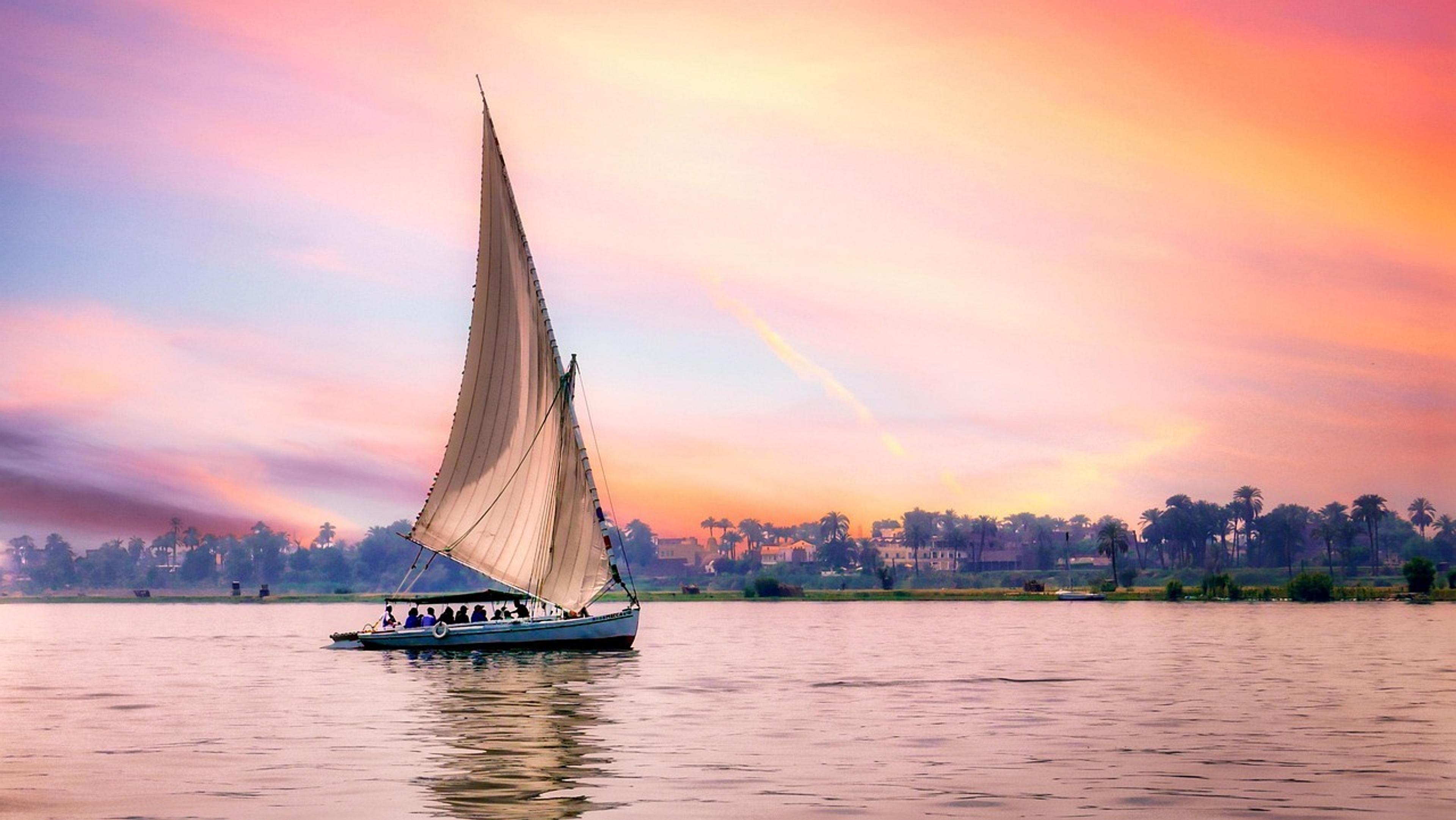 A sailboat glides peacefully along the Nile River at sunset, framed by a vibrant sky and palm trees.