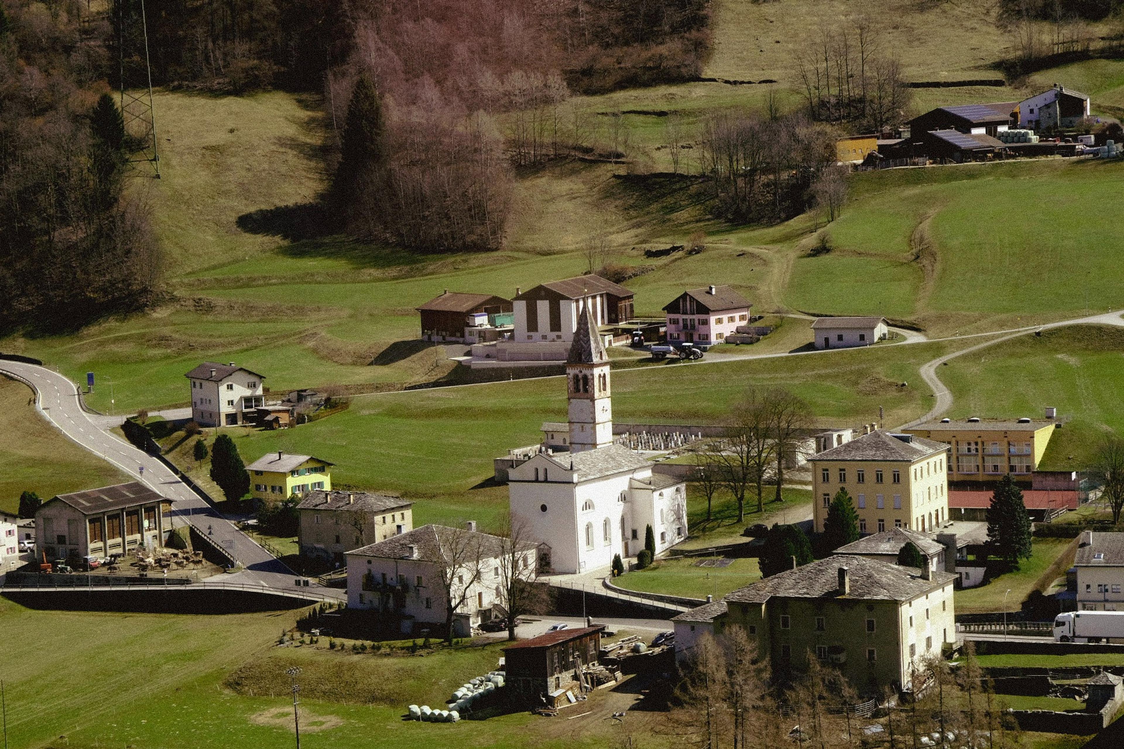 A picturesque view of Santa Maria in Calanca, Switzerland, featuring a prominent church with a tall spire surrounded by a small cluster of houses and lush green hills.