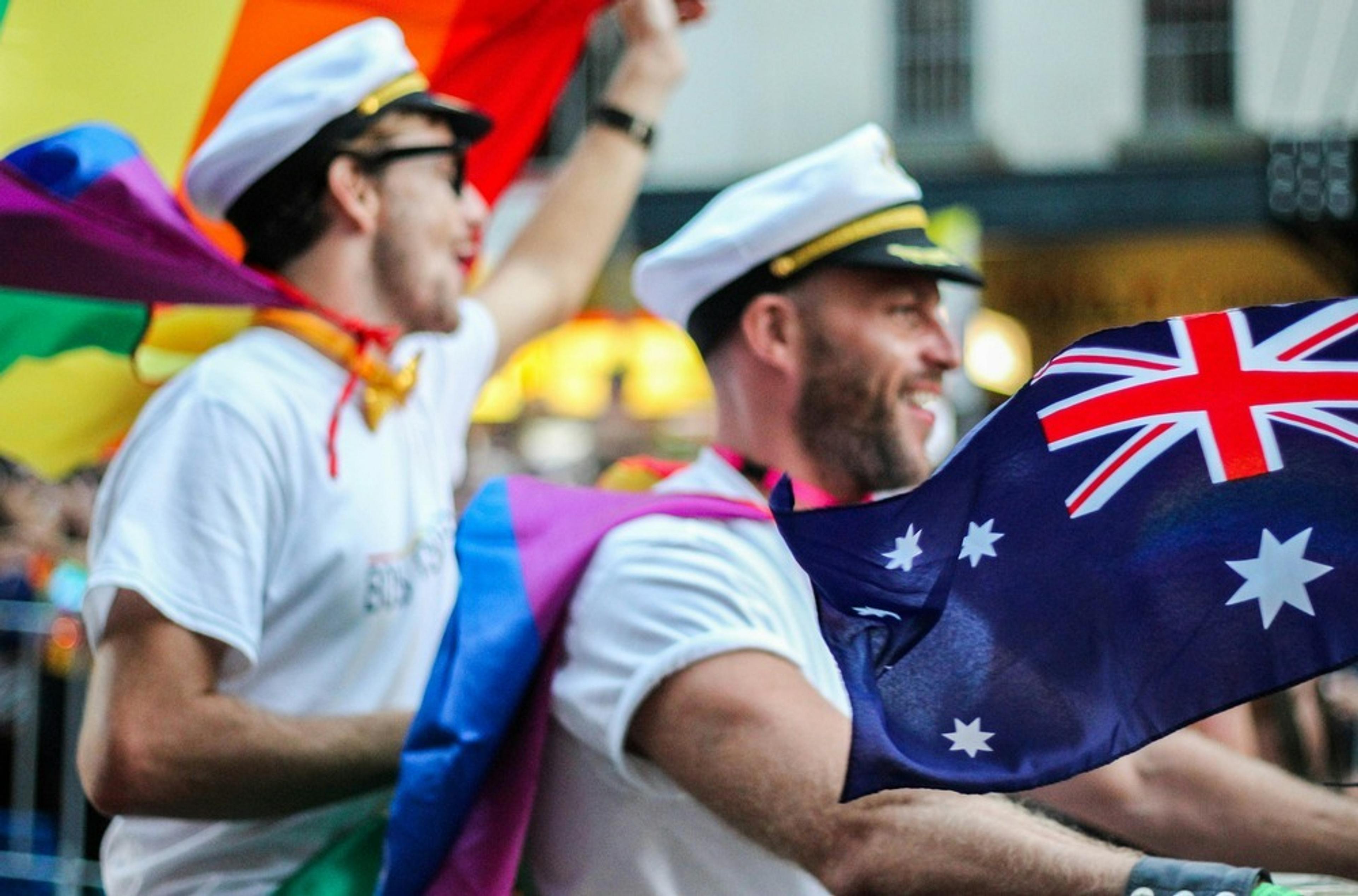 Two individuals in sailor hats joyfully wave rainbow and Australian flags during a parade, possibly at Sydney Mardi Gras.