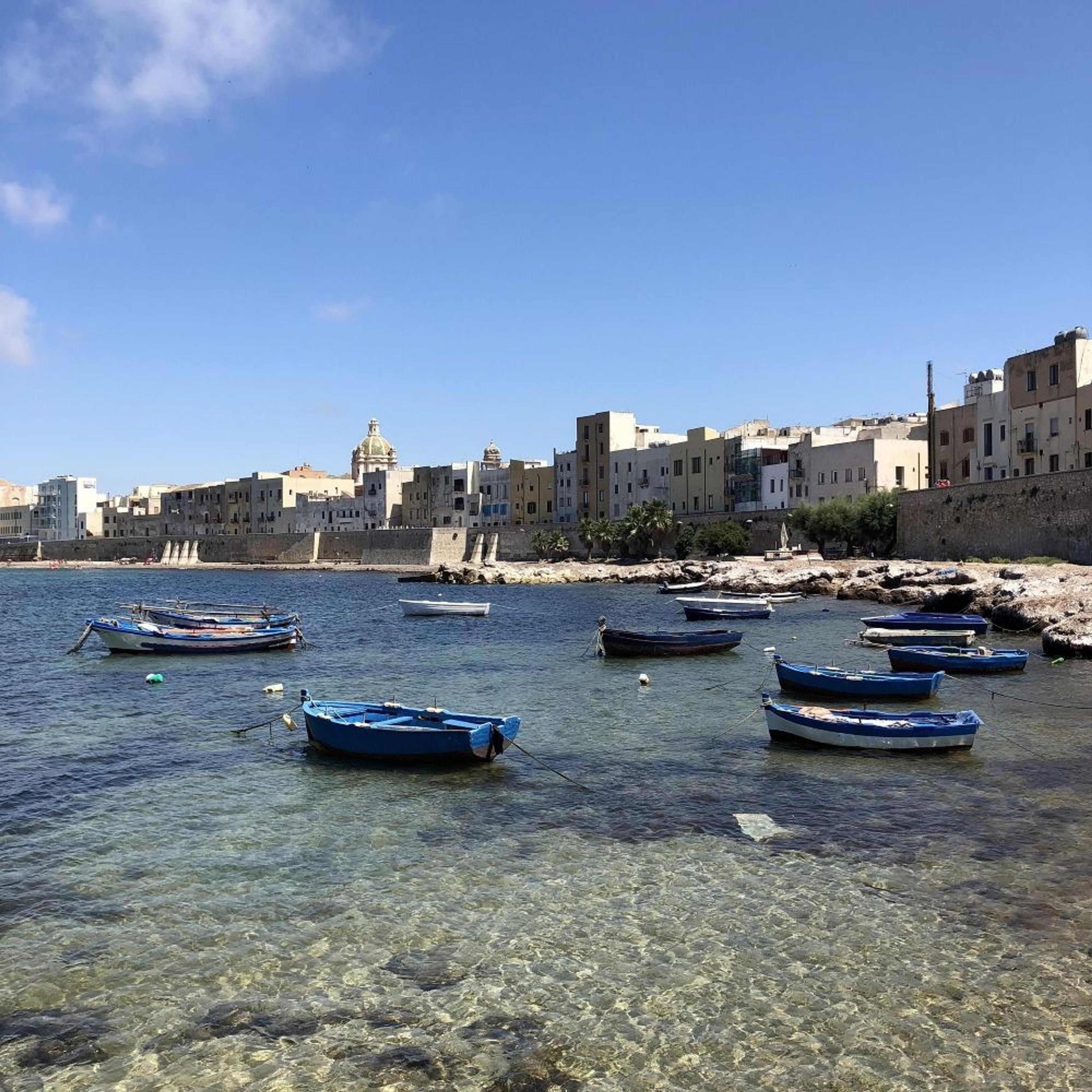 Small blue boats float on crystal-clear waters near the historic coastal buildings of Trapani, Sicily.
