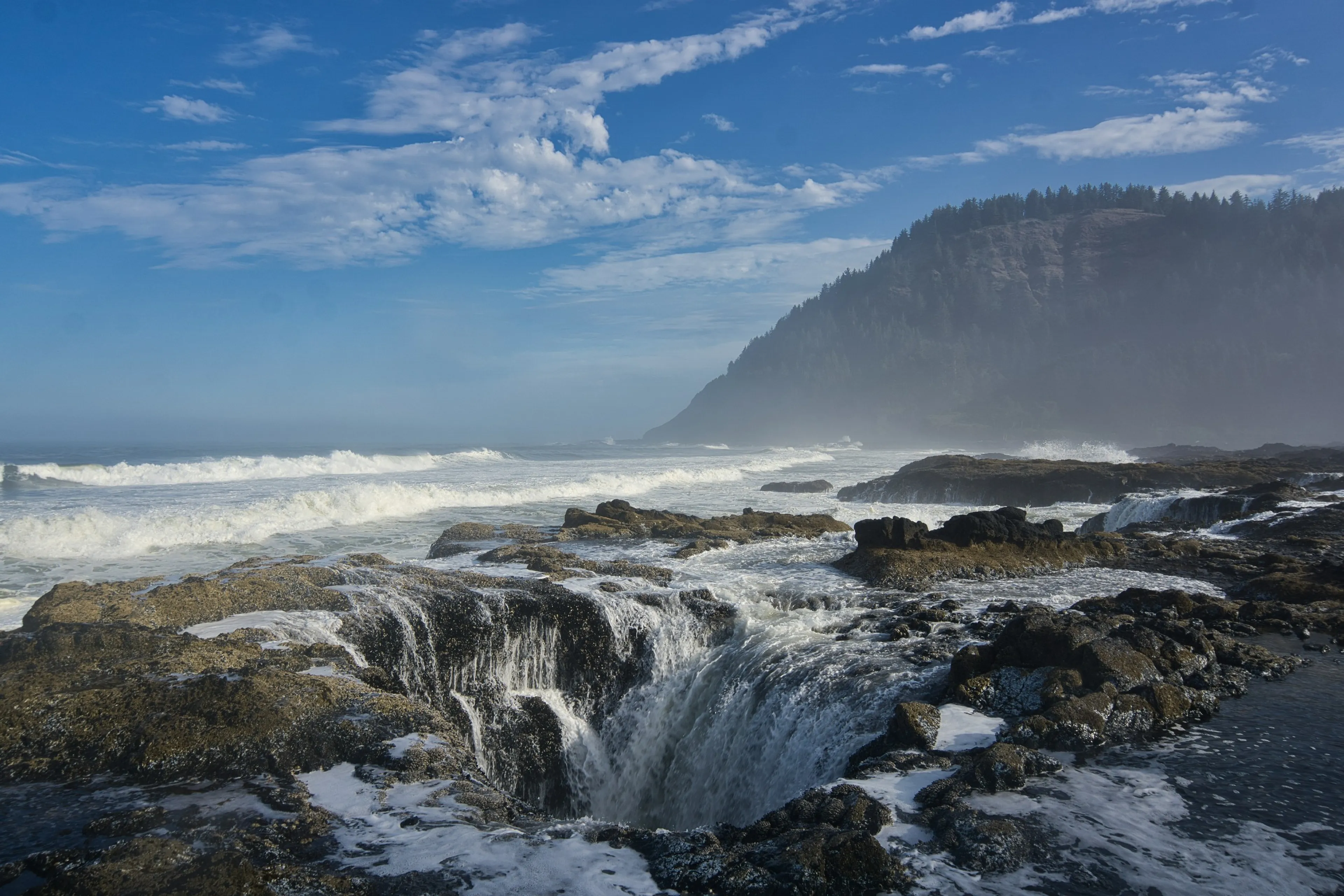 Waves cascade over the rocky Thor's Well along the Oregon coast, with a backdrop of misty hills and a vibrant blue sky.
