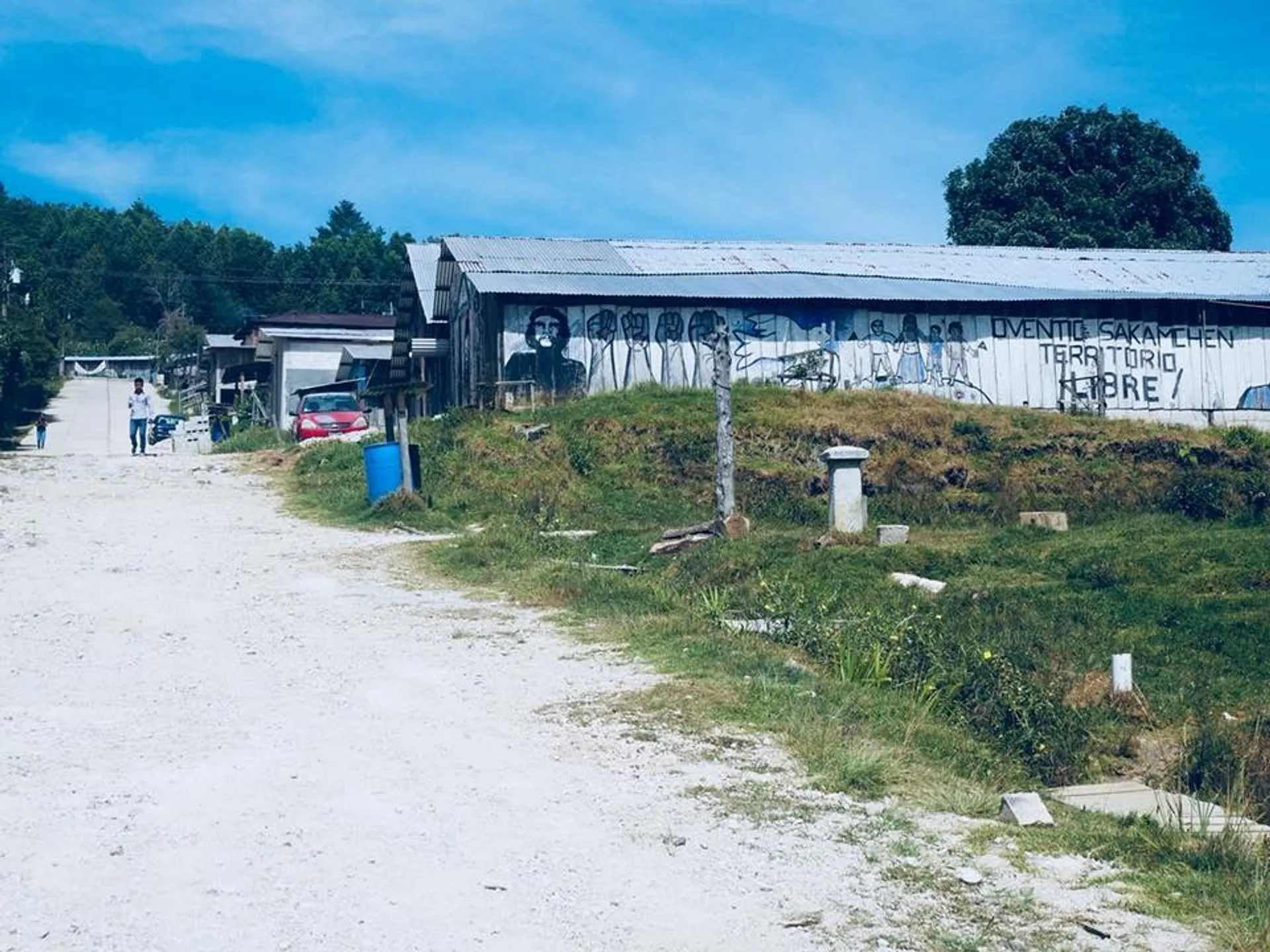 A rural dirt road in Oventic, Chiapas, Mexico, leads to a painted structure featuring murals and text.