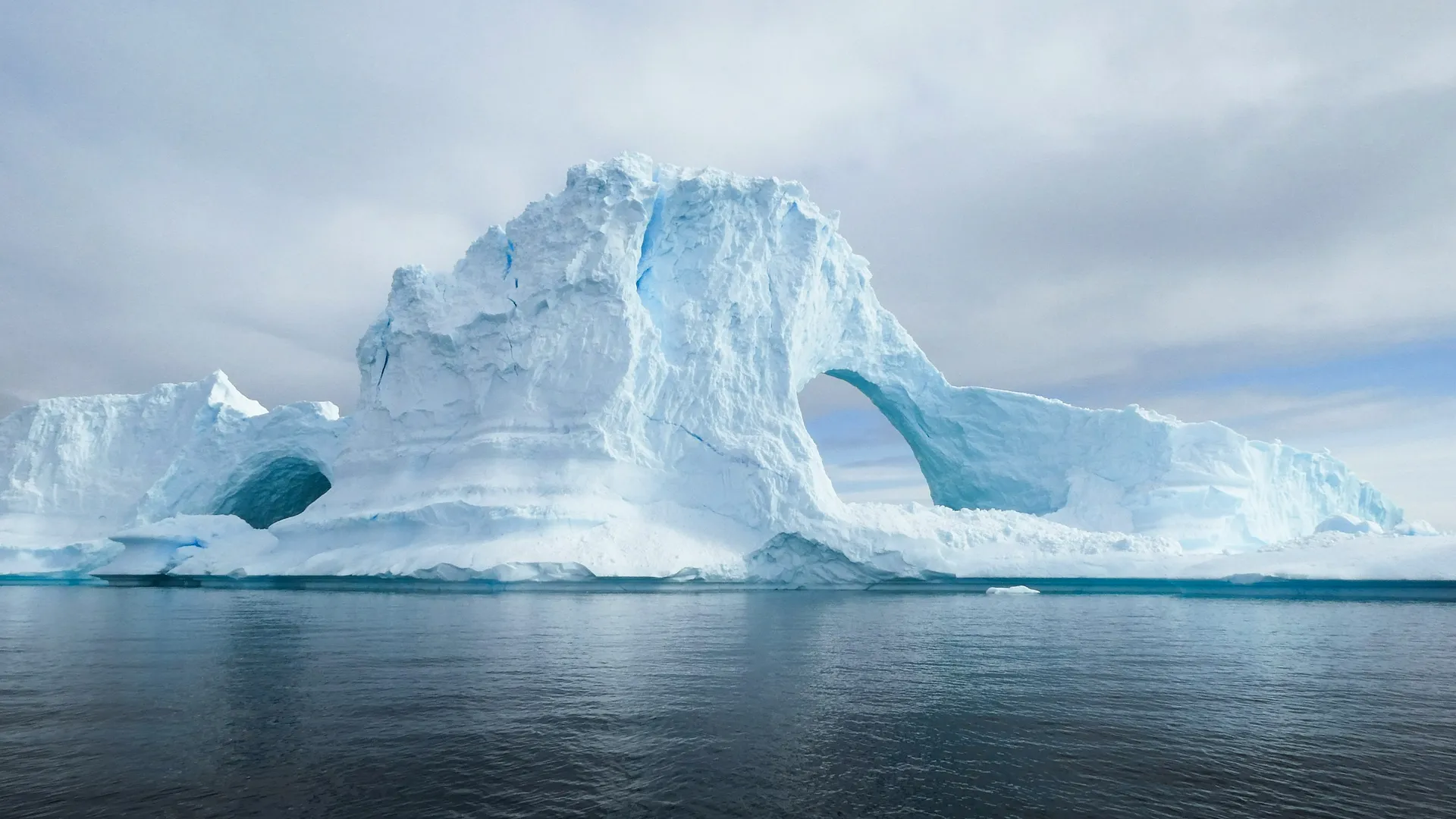 A massive iceberg with natural arches rises majestically from the waters of Antarctica under a cloudy sky.
