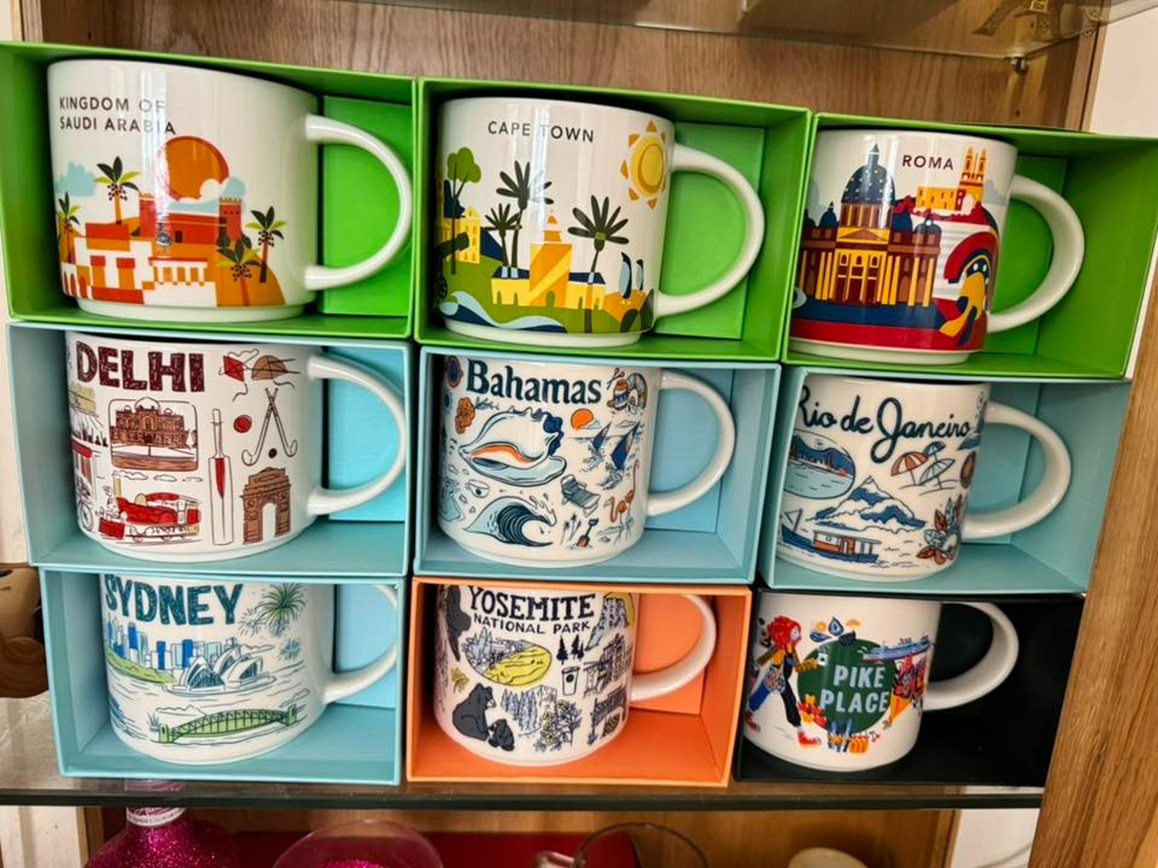 A collection of colorful mugs featuring designs from various destinations, including Cape Town, Roma, and Sydney, is neatly displayed on a shelf.