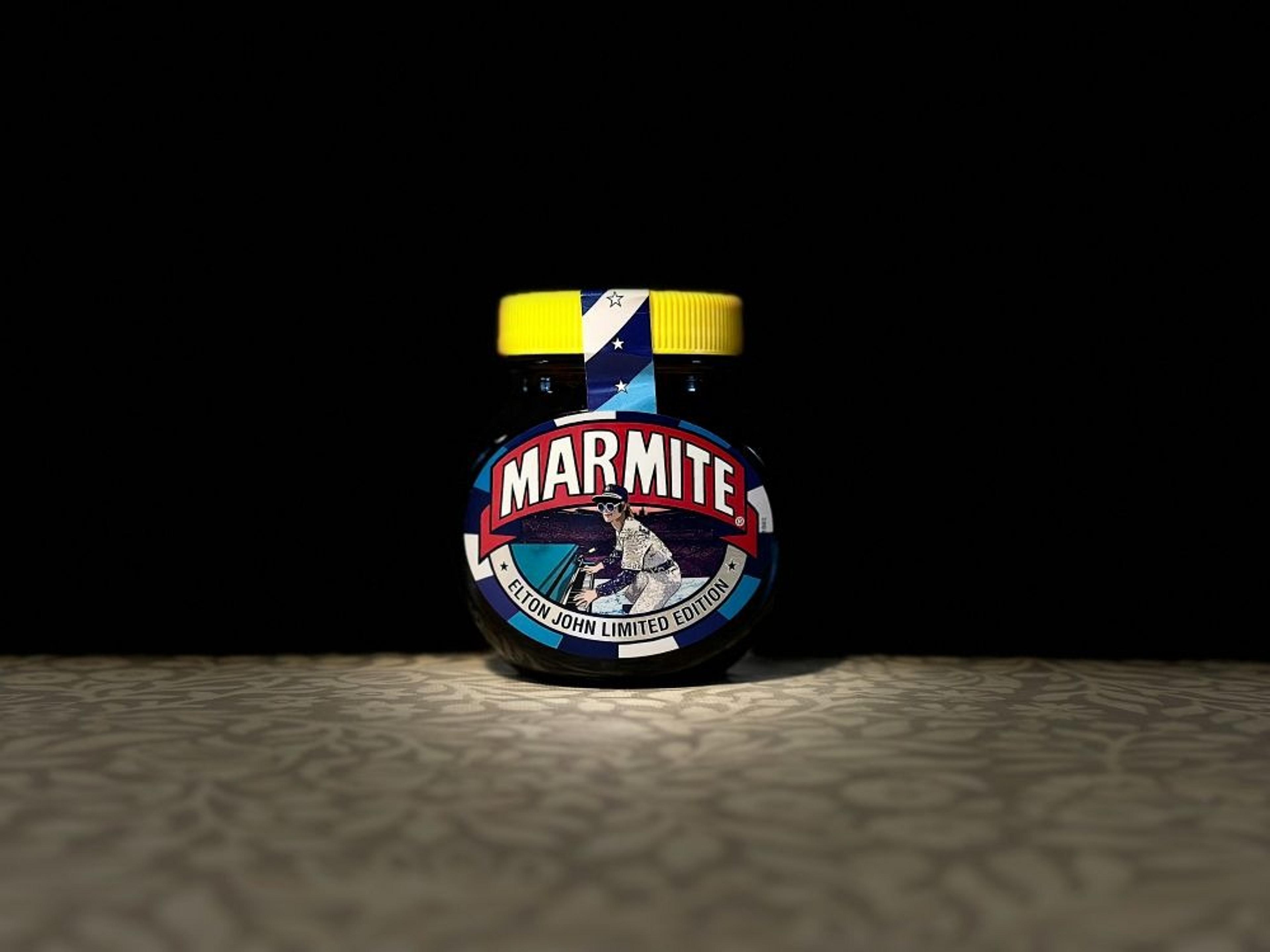 A limited edition jar of Marmite featuring a design inspired by Elton John stands on a patterned surface against a dark background.
