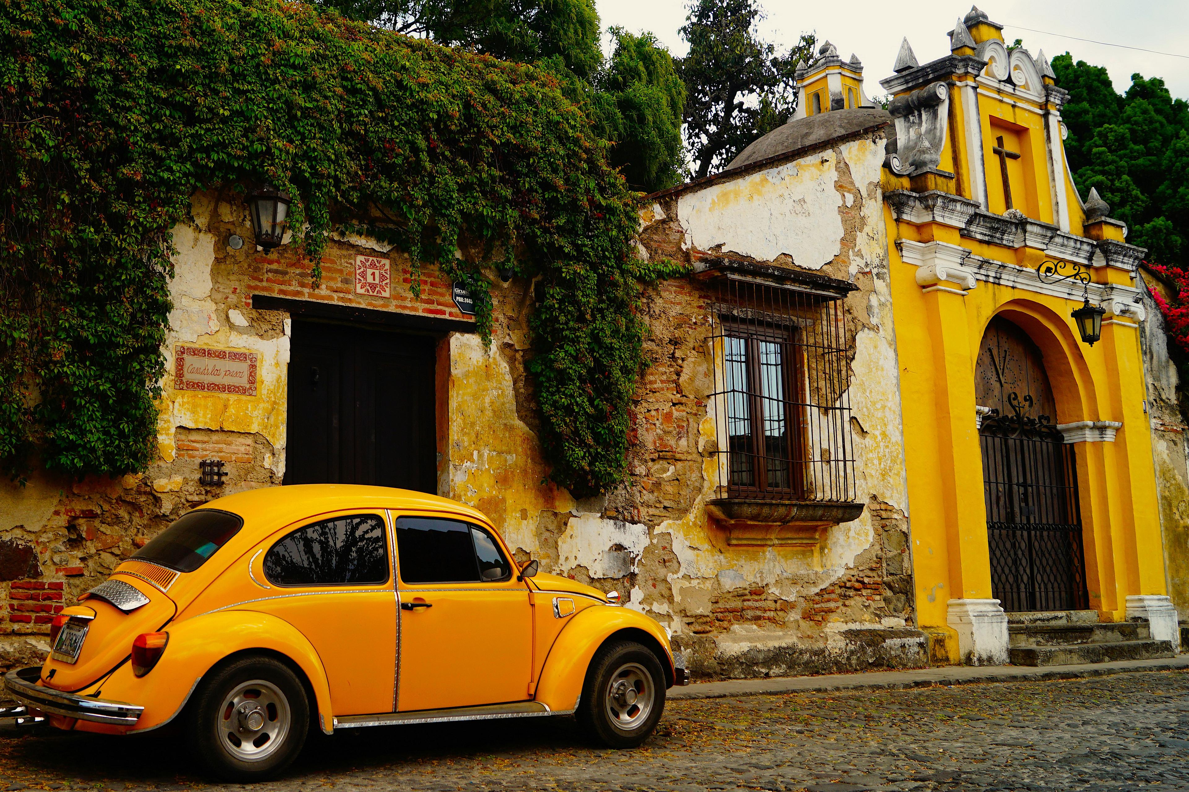 A vibrant yellow Volkswagen Beetle is parked in front of an ivy-covered colonial building in Antigua, Guatemala.
