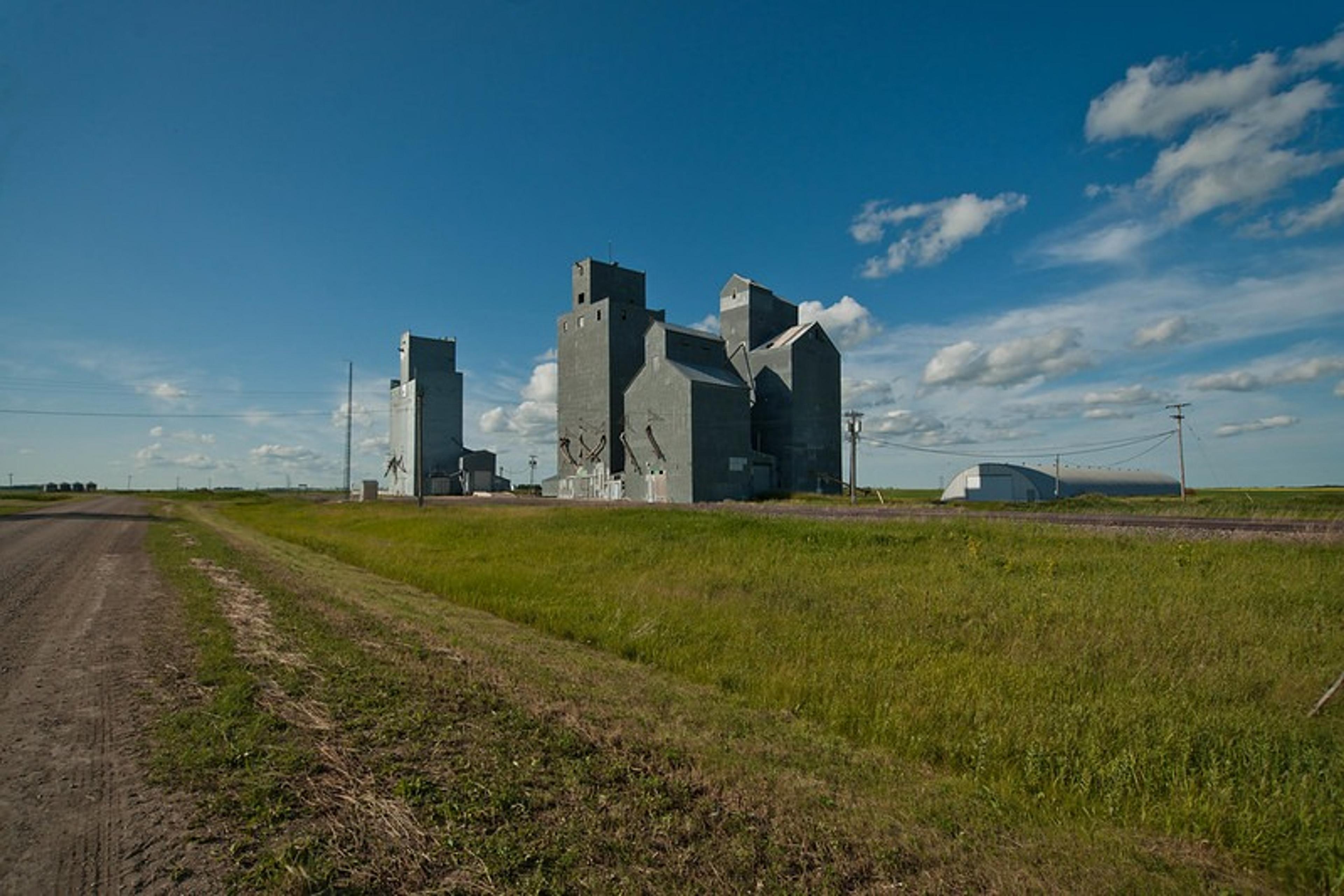 A vast prairie landscape in Saskatchewan features towering, weathered grain elevators under a bright blue sky with scattered clouds.